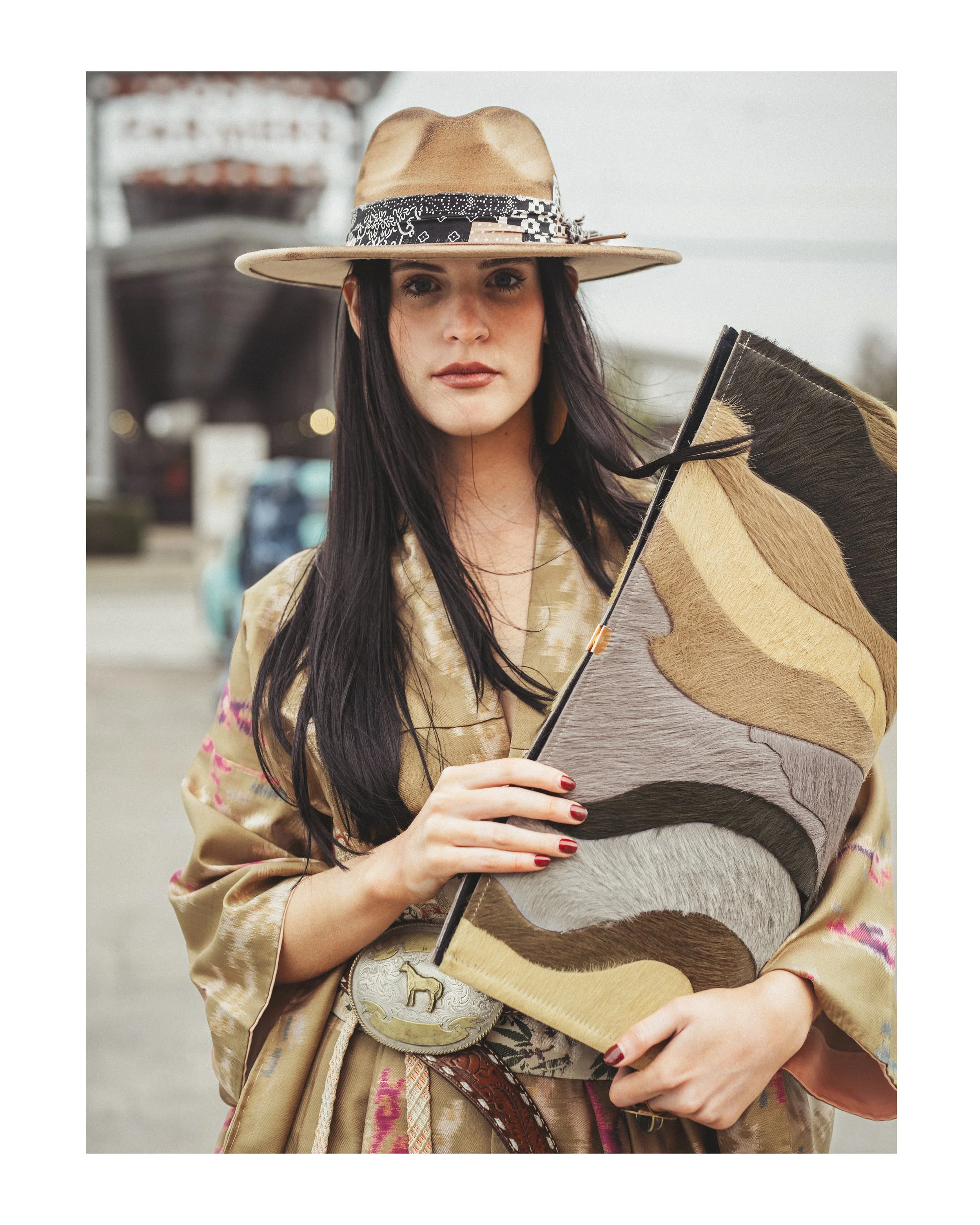 A woman with long dark hair wearing a wide-brimmed hat with a band and traditional clothing, holding a painted canvas with black, brown, and beige horns.