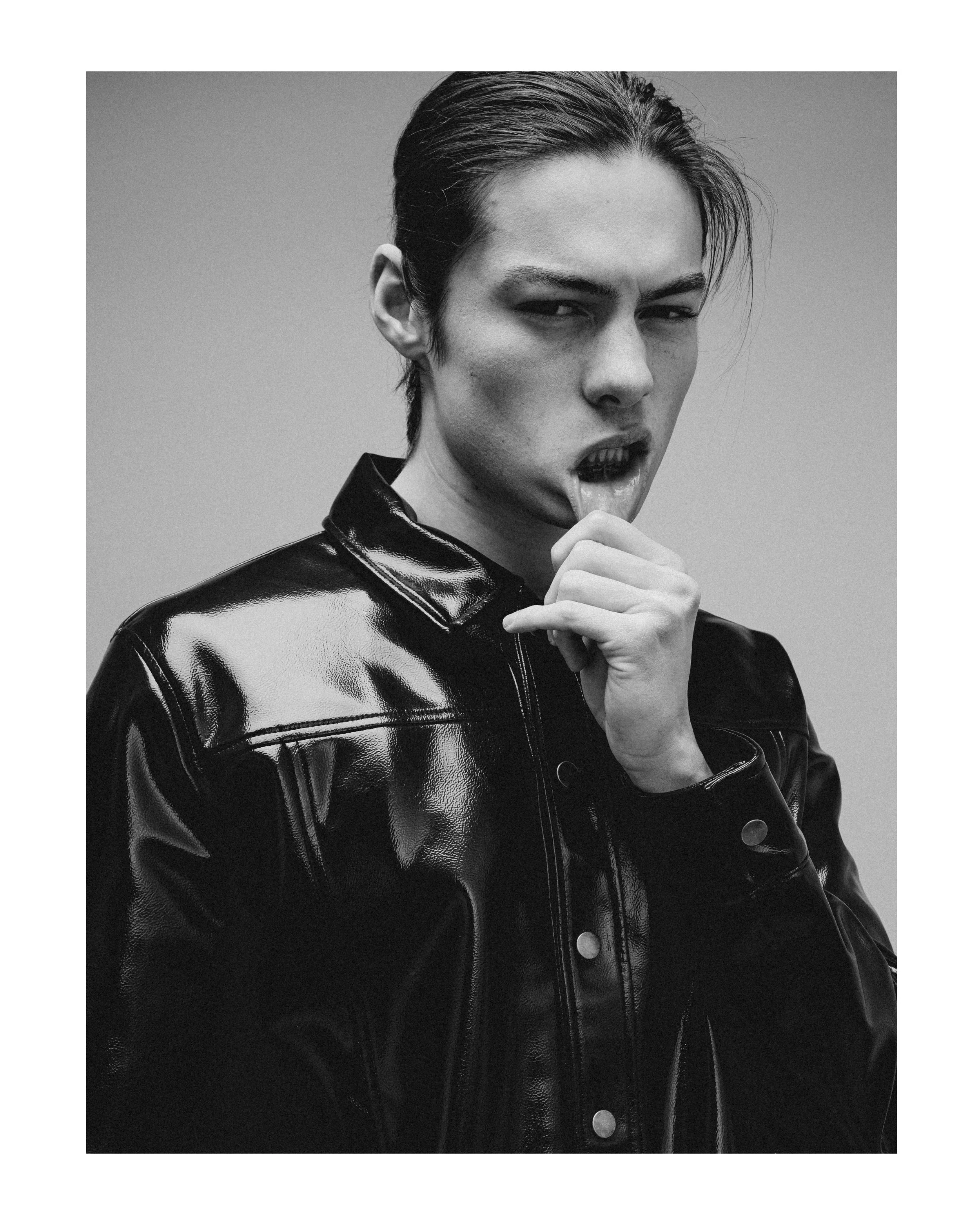 A person with dark hair slicked back, wearing a shiny black leather jacket, sticking out their tongue and holding their chin with one hand in a confident pose.