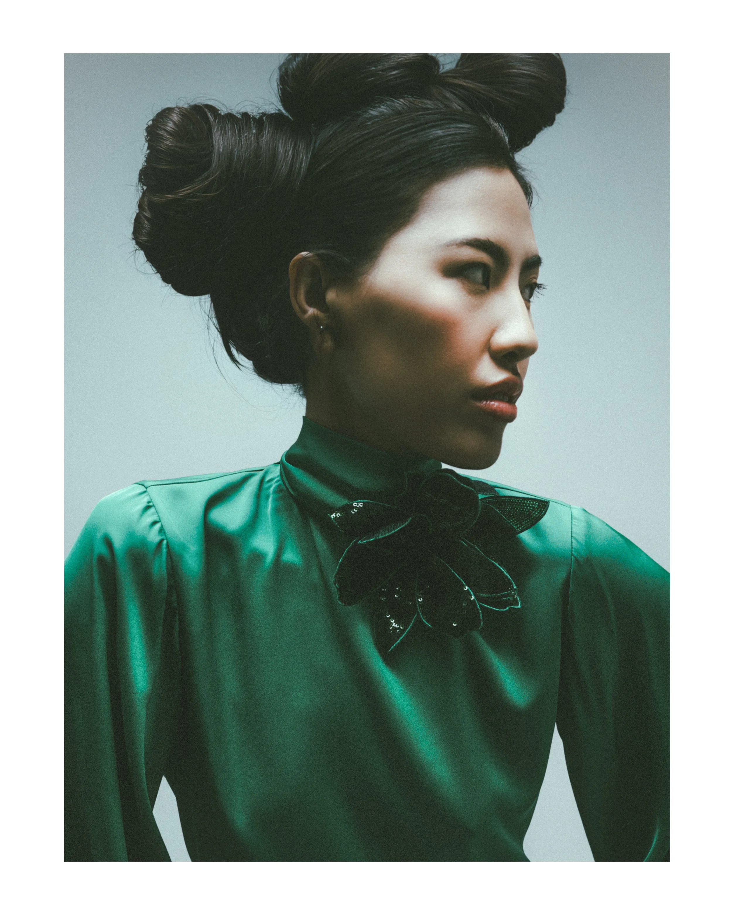 Close-up of a woman with dark hair styled in an elaborate updo, wearing dark makeup and a green satin outfit with a black bow or ribbon at the collar, against a plain background.