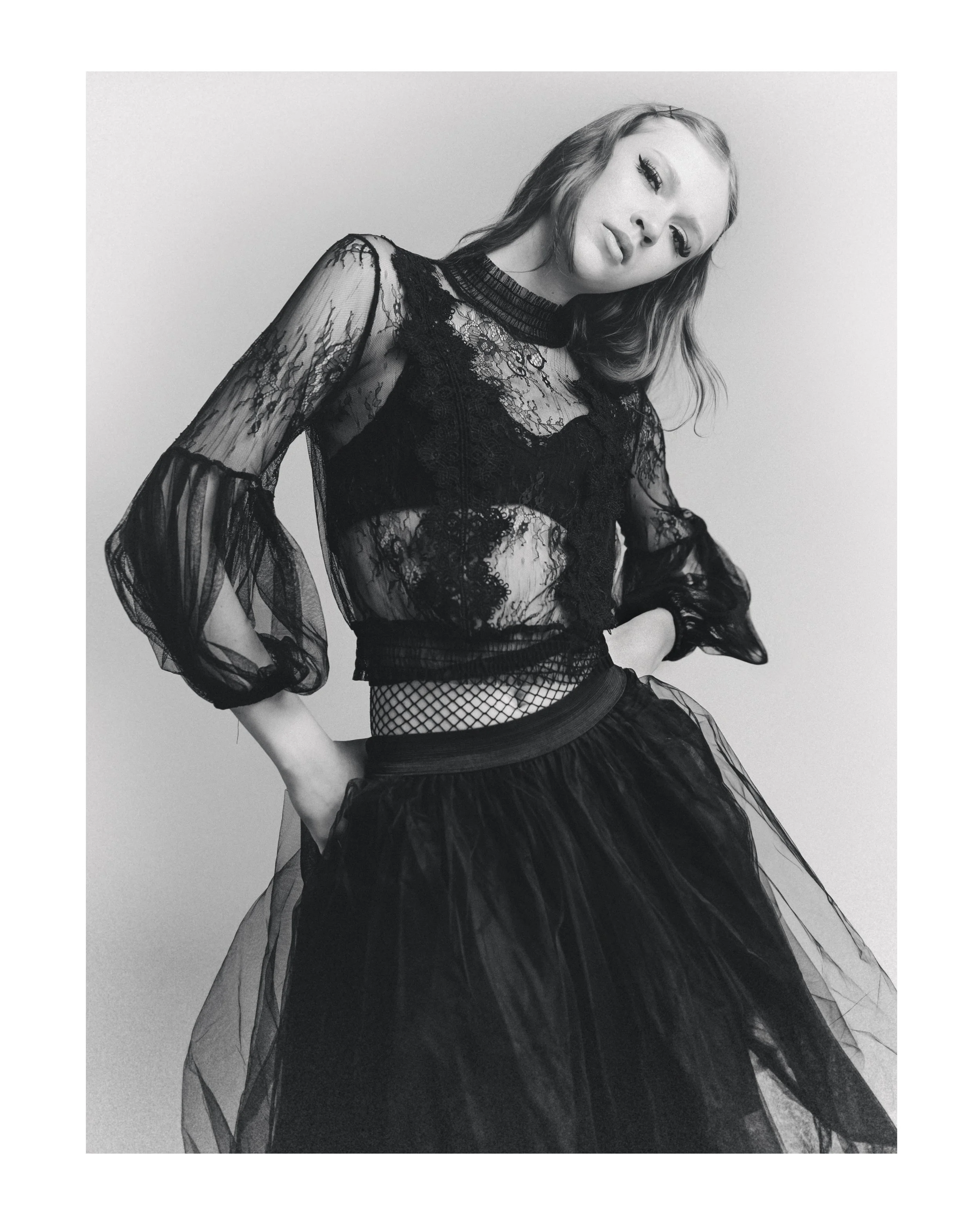 A woman with wavy hair wearing a black lace top with sheer sleeves and a layered black skirt, posing with her head tilted and serious expression in a studio with a plain backdrop.