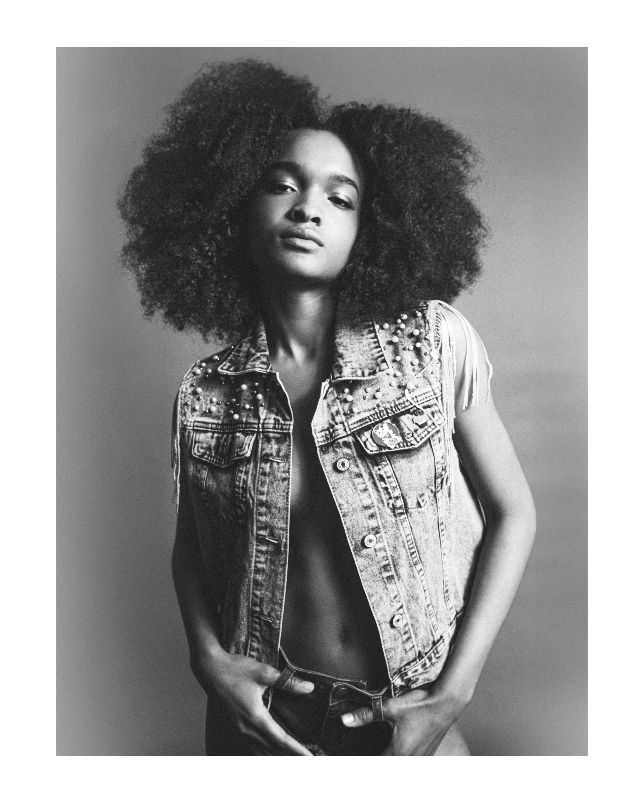 Black and white photograph of a woman with voluminous afro hairstyle, wearing a studded denim vest and a leather belt, standing against a plain background.