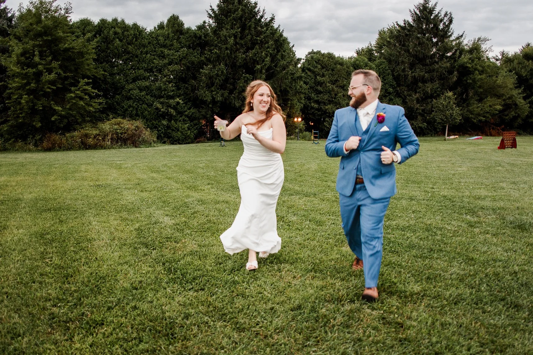 wide shot of bride and groom running at camera, laughing and looking at each other with drinks in hand in field at bloom and bower in rising sun ohio