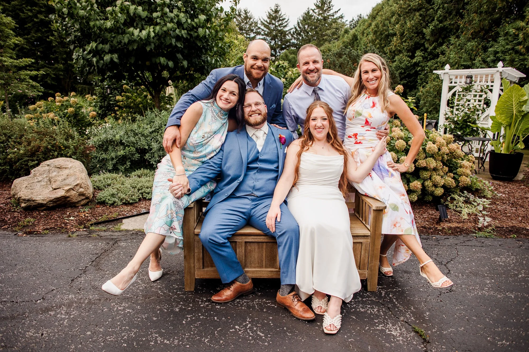bride and groom sitting on bench with a few wedding guests surrounding them and smiling at the camera