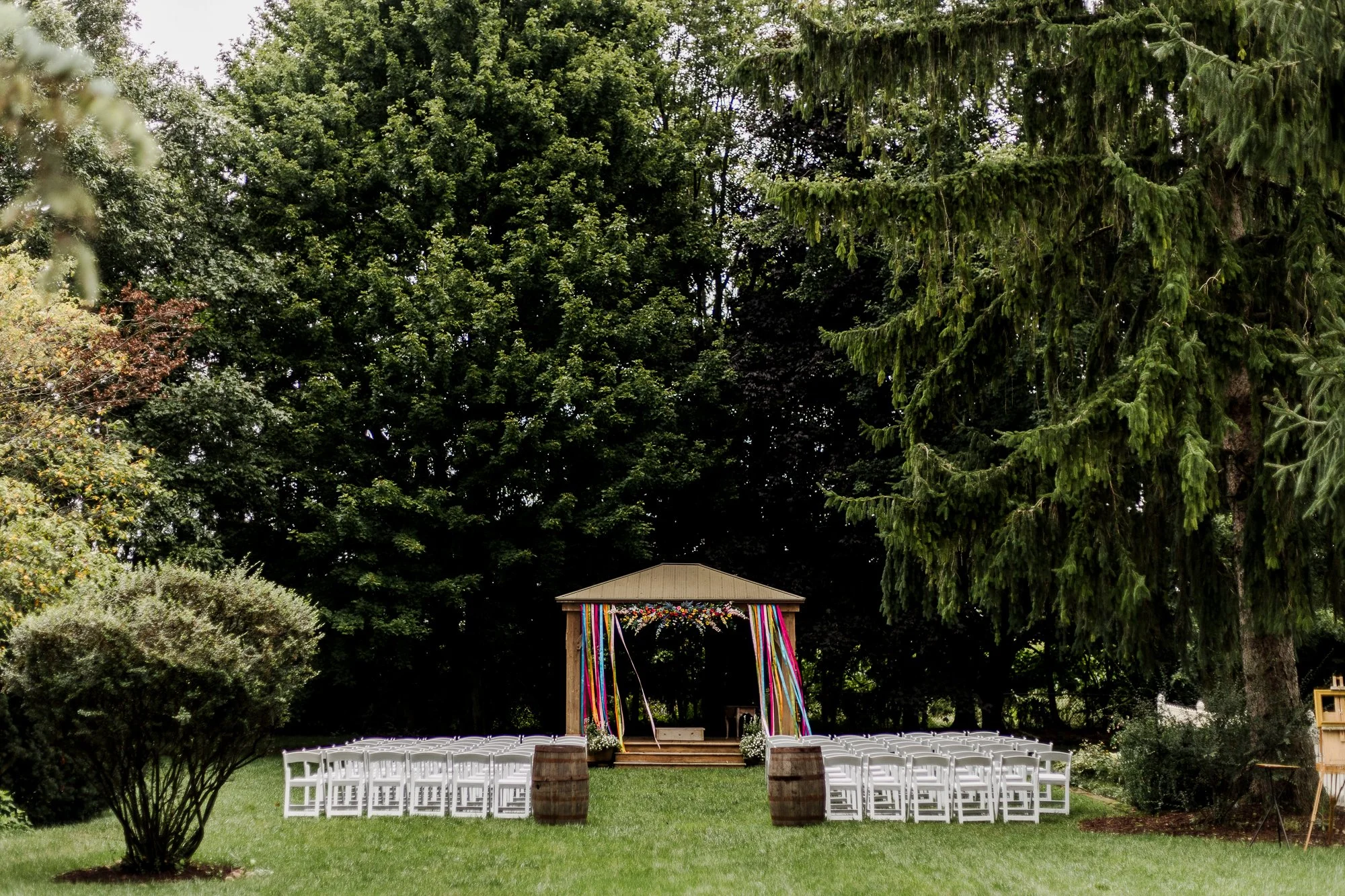 Ceremony site in beautiful green lawn has a gazebo with rainbow colored ribbons hanging and blowing in the wind