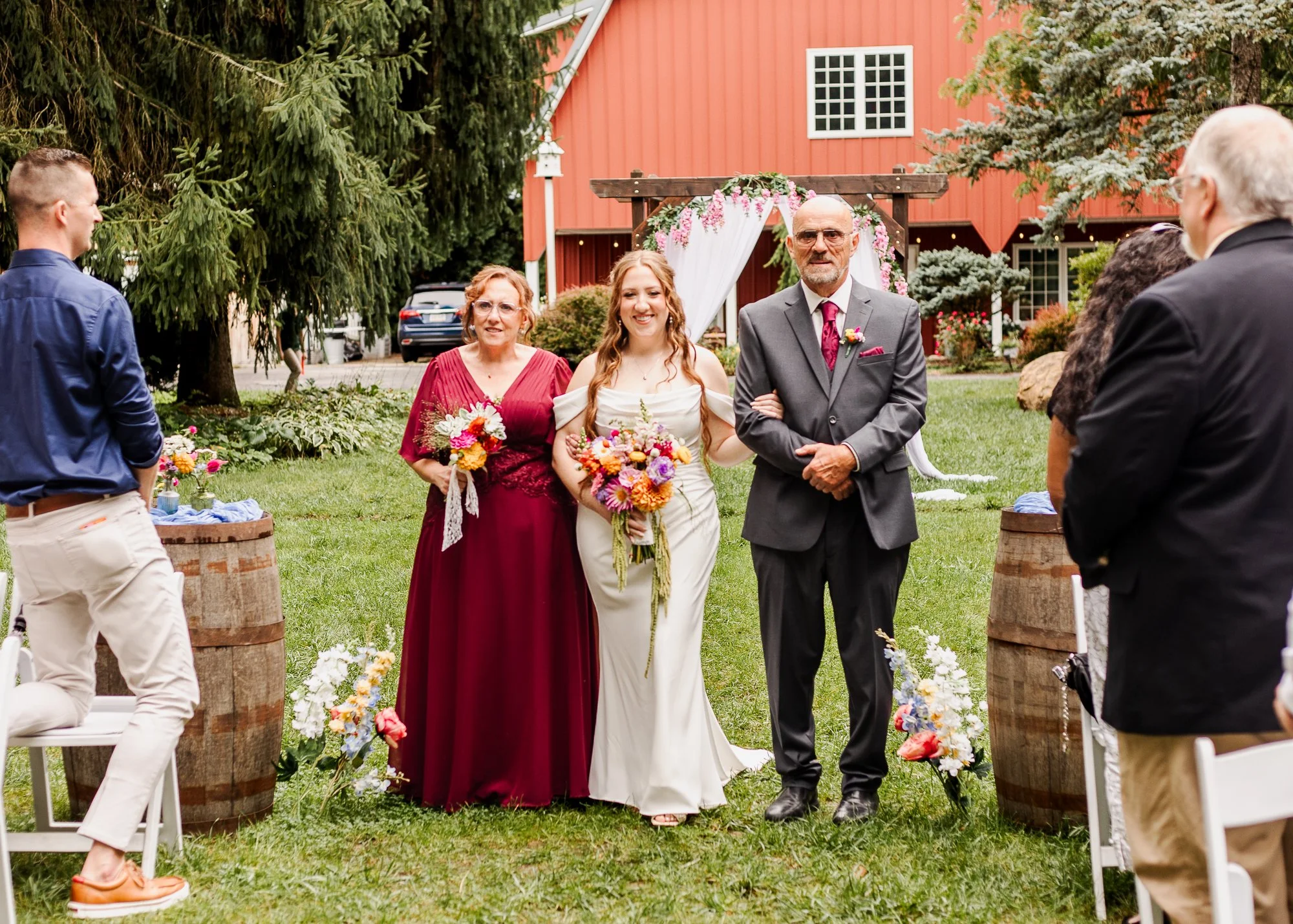 Bride walks down aisle with her mom and dad during her ceremony at bloom and bower in rising sun ohio with red barn in background