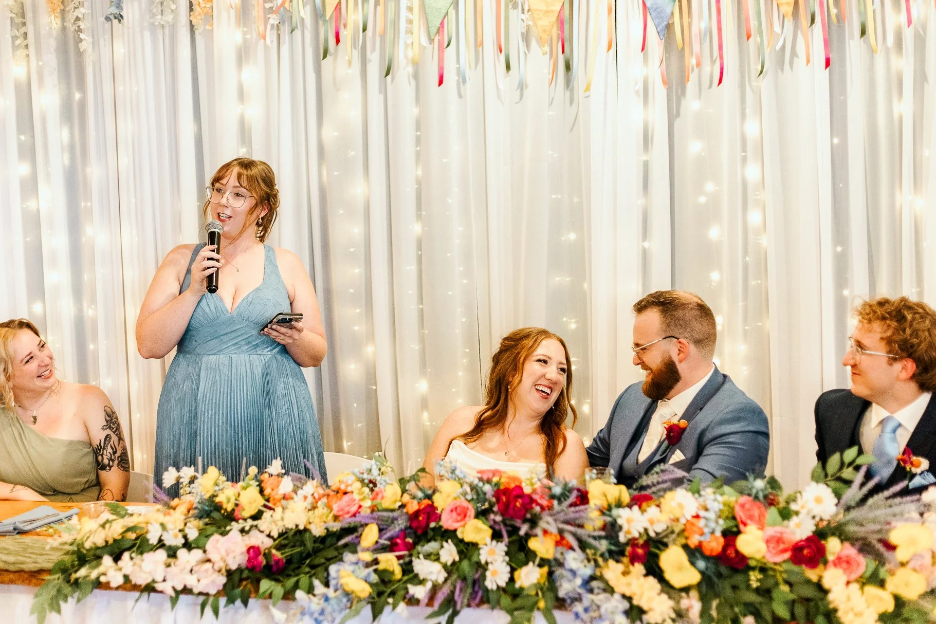 wide shot of sister of the bride toasting to the bride and groom at head table while bride and groom look at each other, laughing