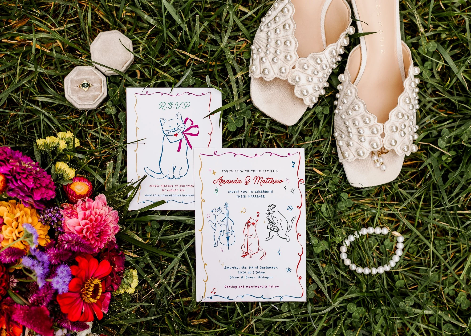 Flatlay of bridal details including pearl adorned shoes, emerald engagement ring, cat-themed wedding invitation, and rainbow-colored bouquet