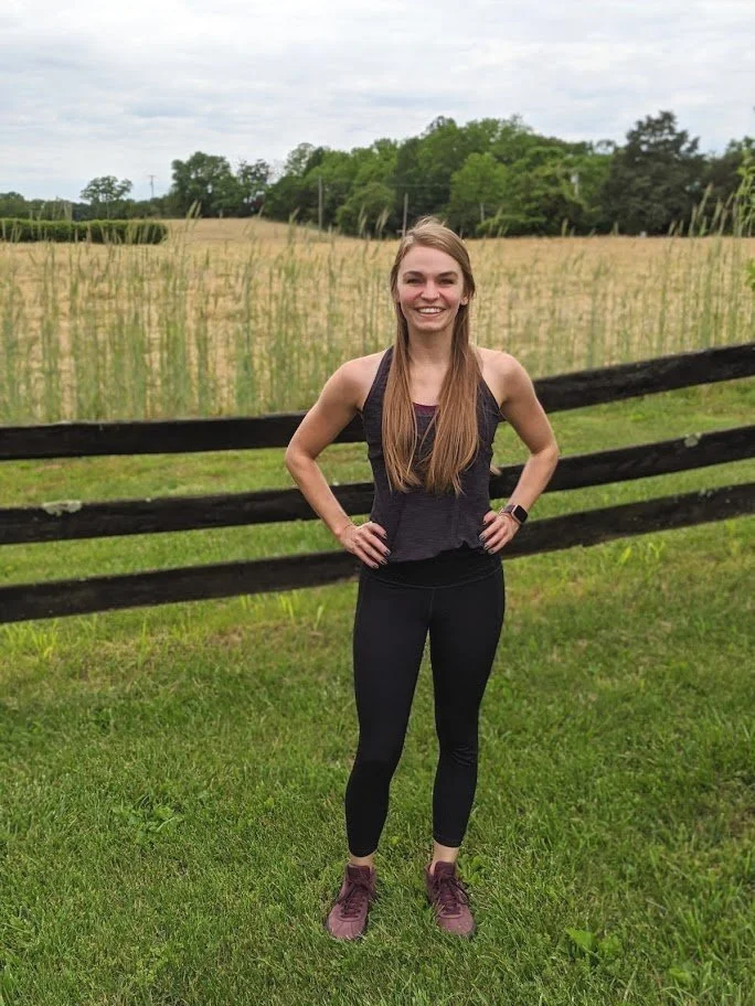 Stephanie Hayslip, the owner of Vitality by Stephanie, stands in front of a green and tan field. She is smiling at the camera and is dressed in black workout clothes and red sneakers.