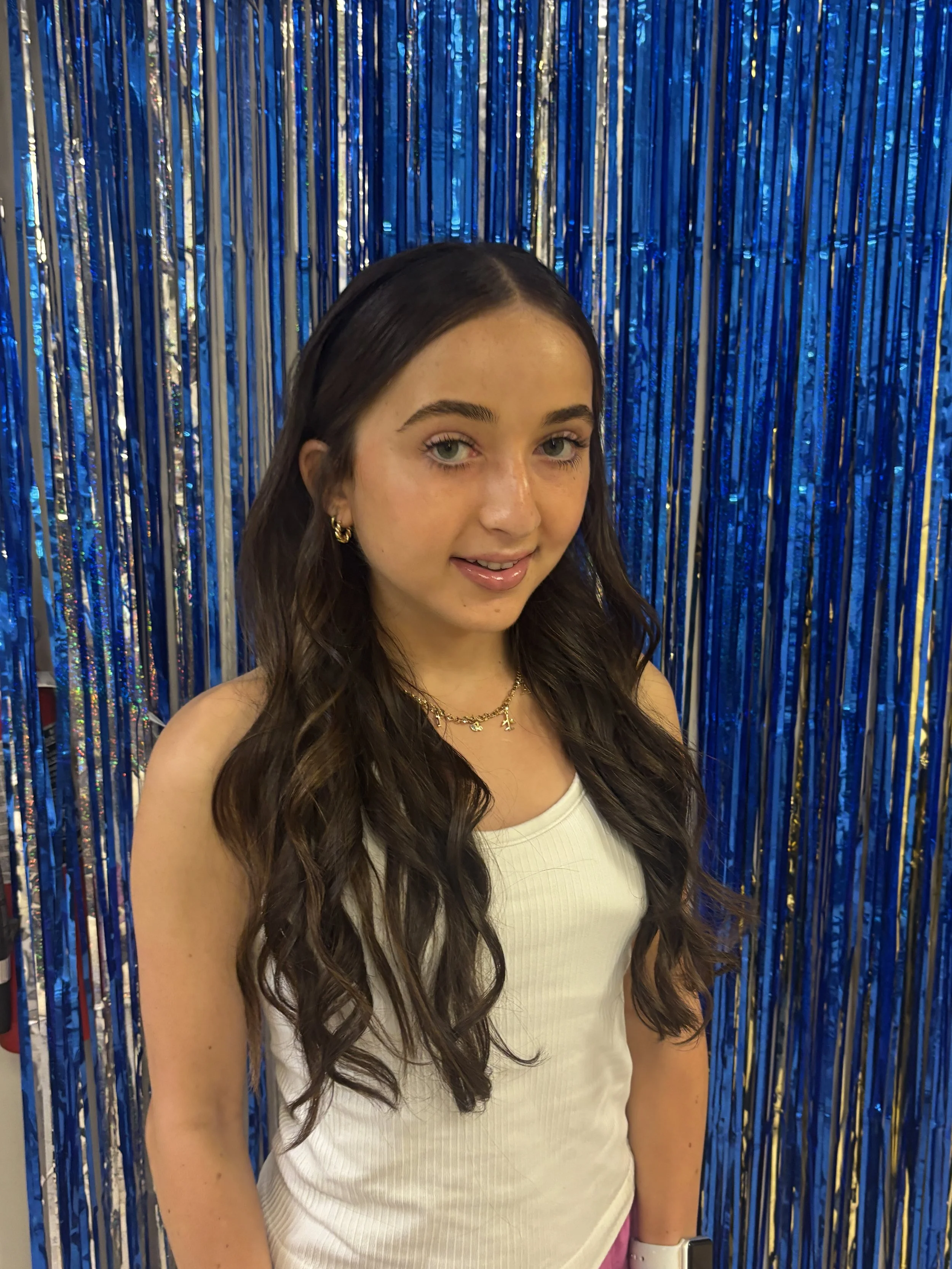 A young woman with long wavy hair, wearing gold earrings, a gold necklace, a white tank top, and pink pants, standing in front of a shiny blue and silver tinsel backdrop.