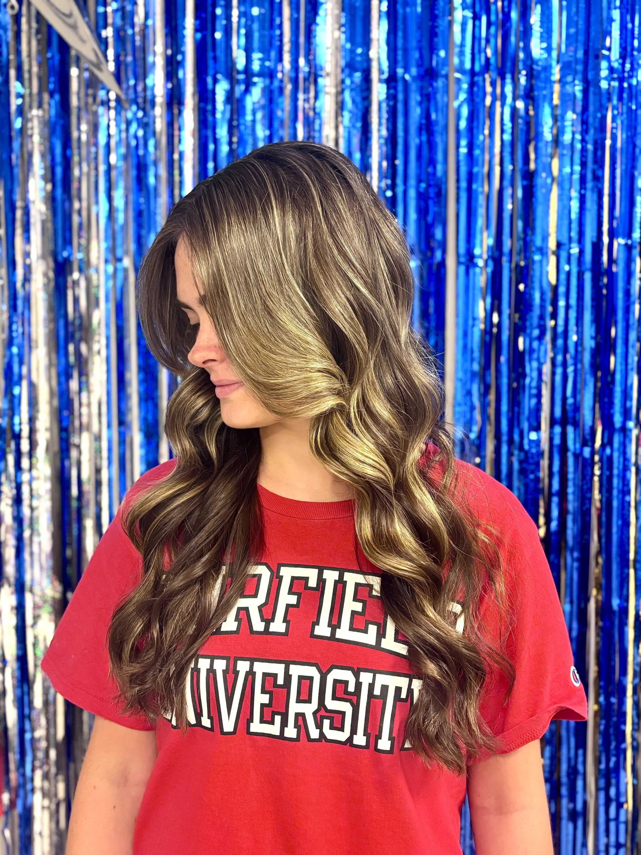 A woman with long, wavy, light brown hair featuring highlights, wearing a red shirt with text. She is standing in front of a blue and silver tinsel backdrop, looking downward with her eyes closed.