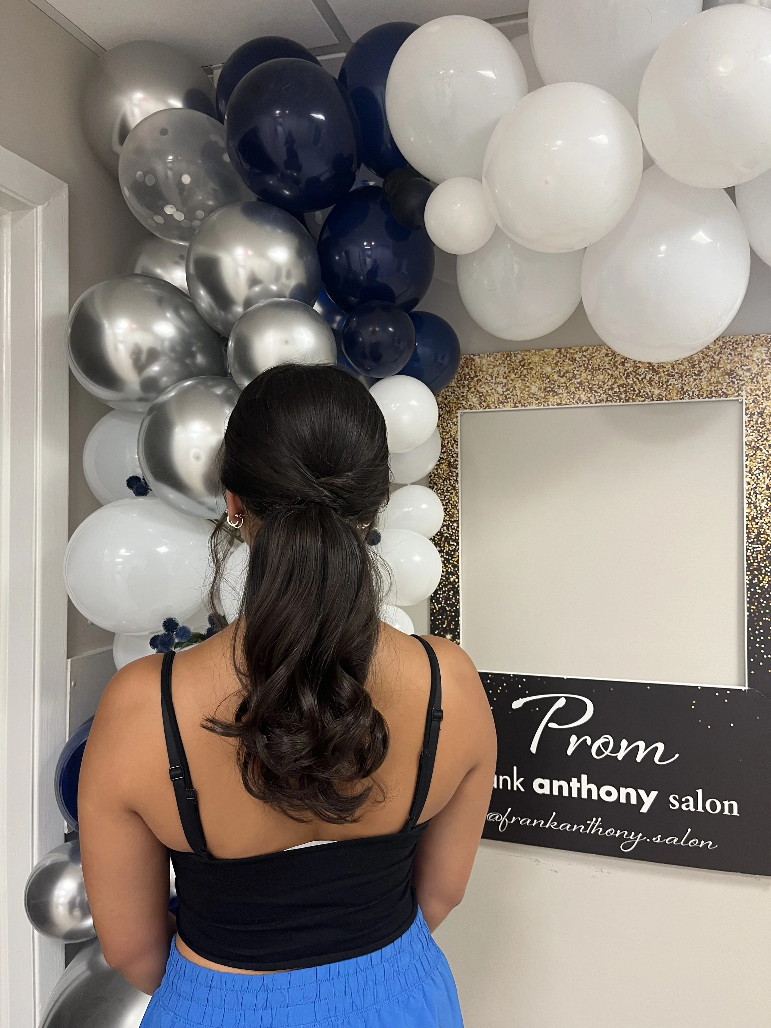 Woman with long dark hair in a black tank top and blue shorts facing away from camera, standing in front of a black board with white lettering and a balloon display with black, silver, white, and transparent balloons.