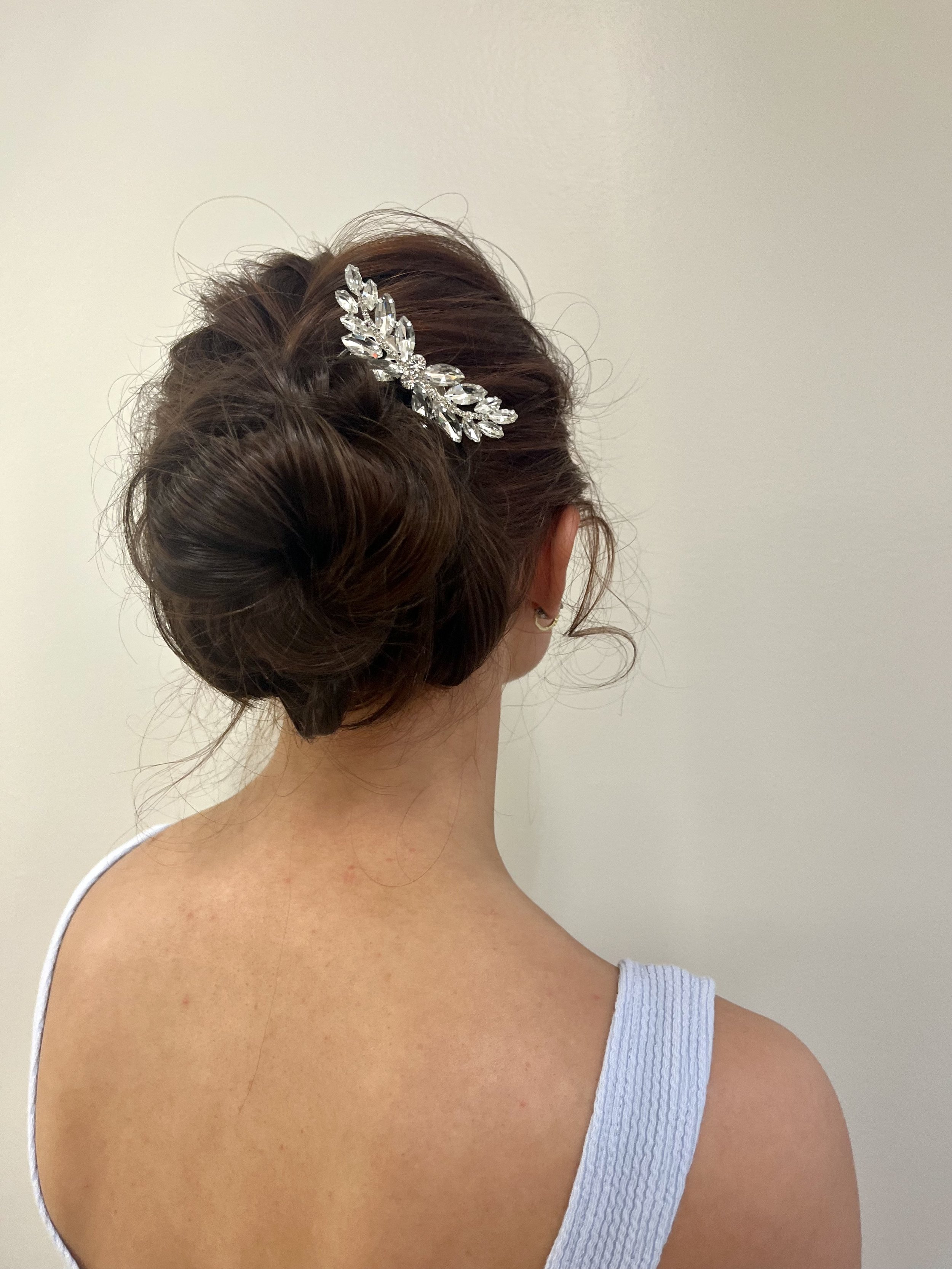 Back view of a woman with an elegant updo hairstyle decorated with a silver and clear crystal hair accessory, wearing a light blue tank top, against a plain light-colored wall.
