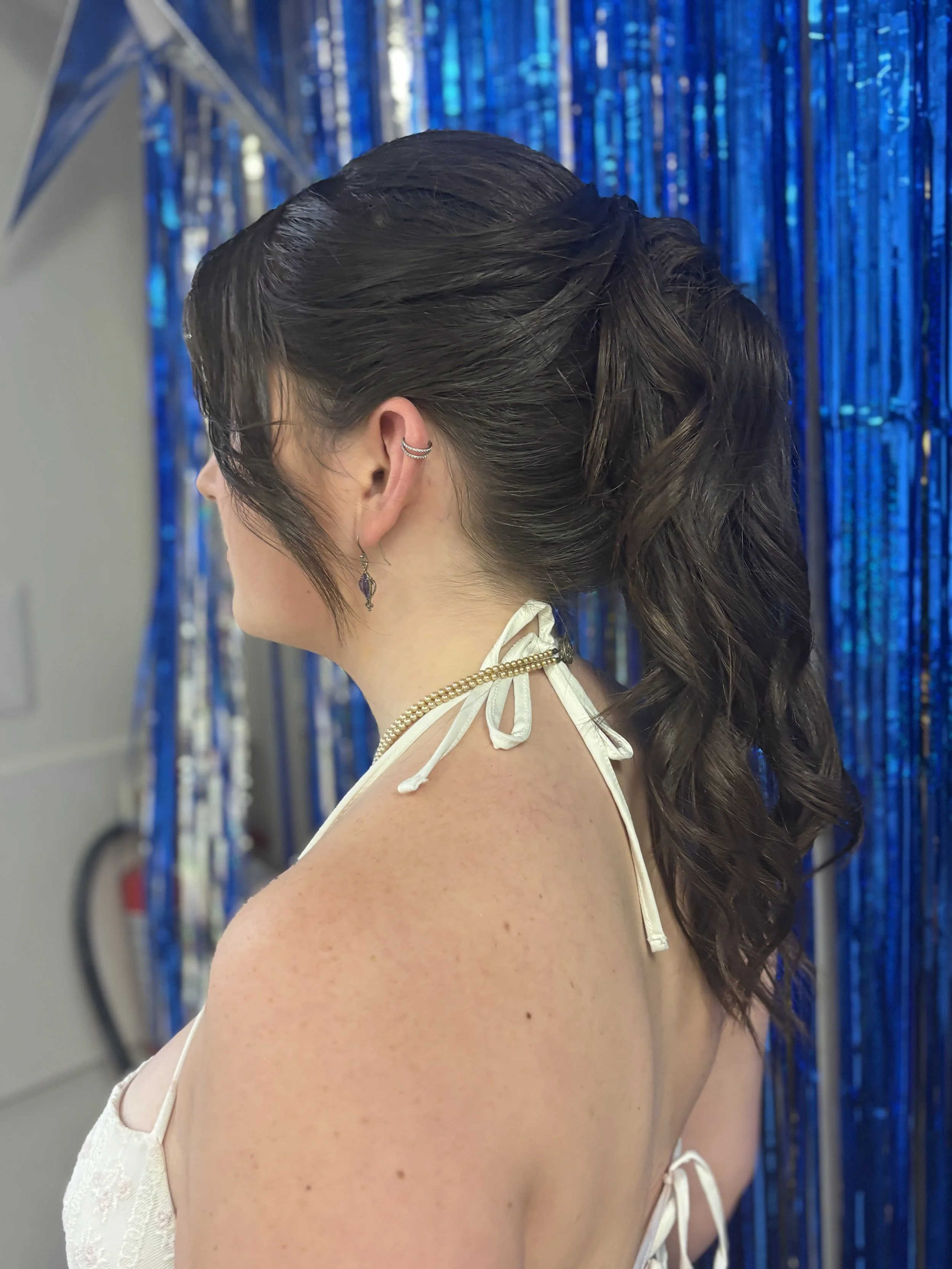 Side profile of a woman with dark brown hair styled in loose curls, wearing earrings and a cream-colored dress with thin straps, standing in front of a blue metallic fringe curtain.