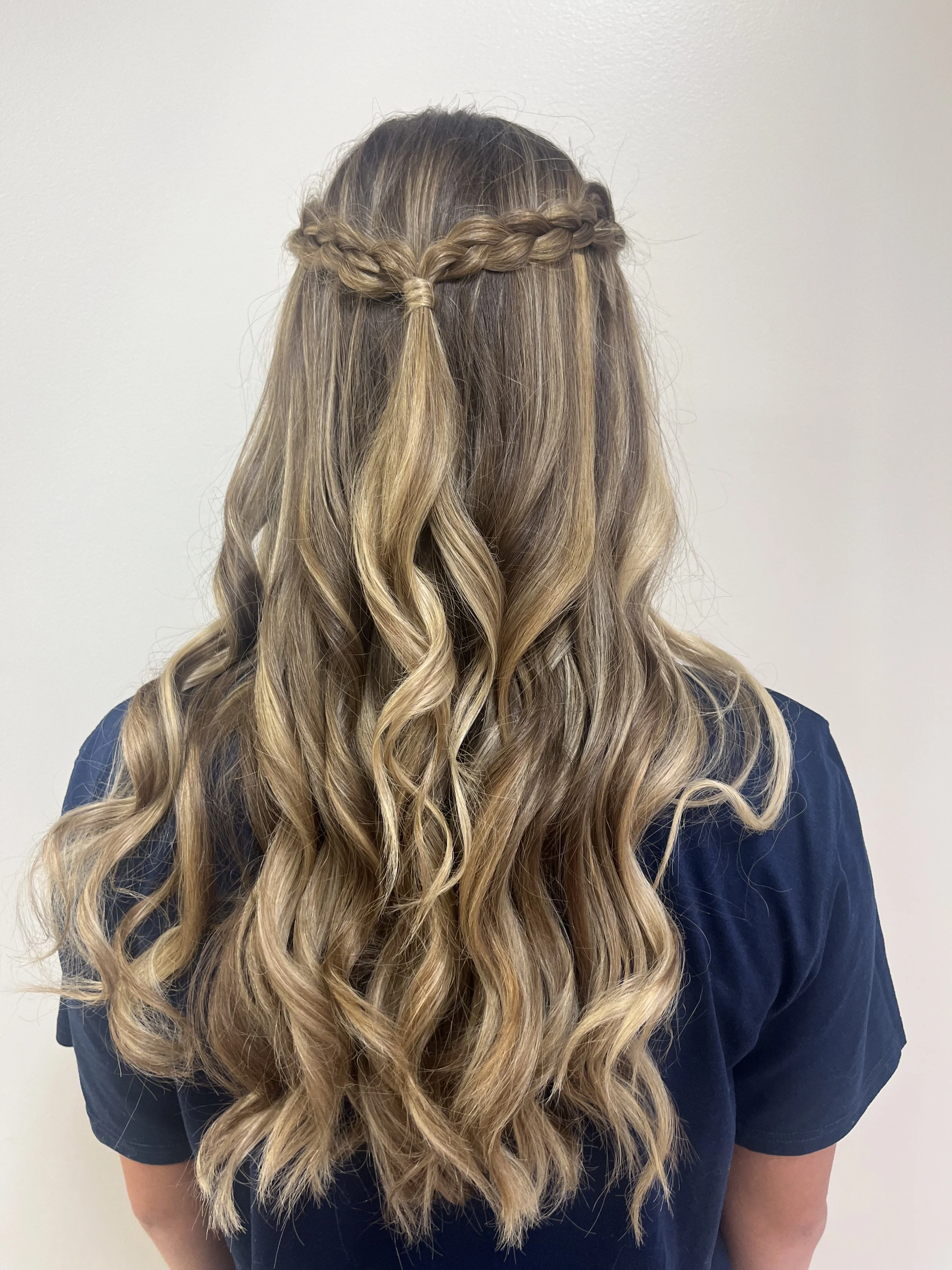 Back view of a woman with long, wavy blonde hair styled with a braid crown and loose curls, wearing a navy blue shirt.