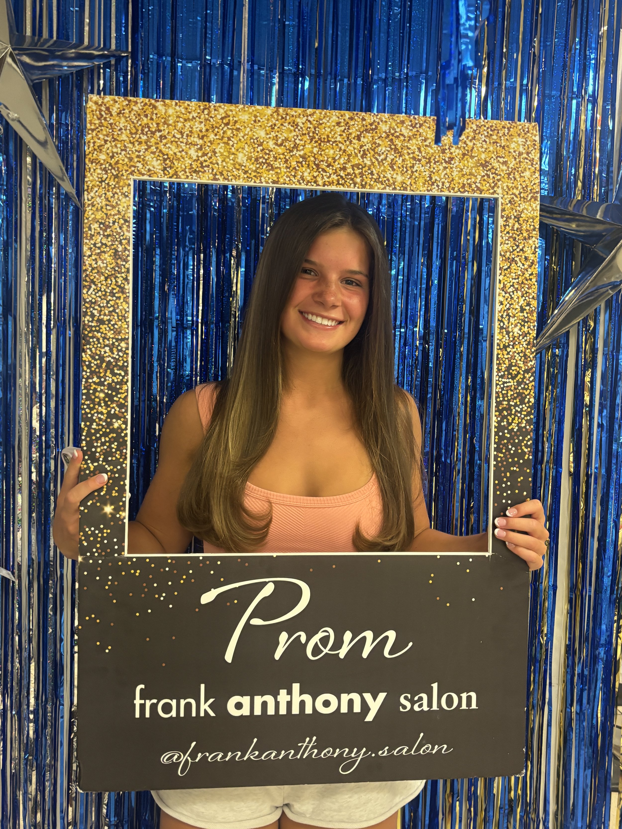 A young woman with long brown hair smiling, holding a black sign that reads 'Prom' in front of a blue and gold tinsel backdrop at Frank Anthony Salon.