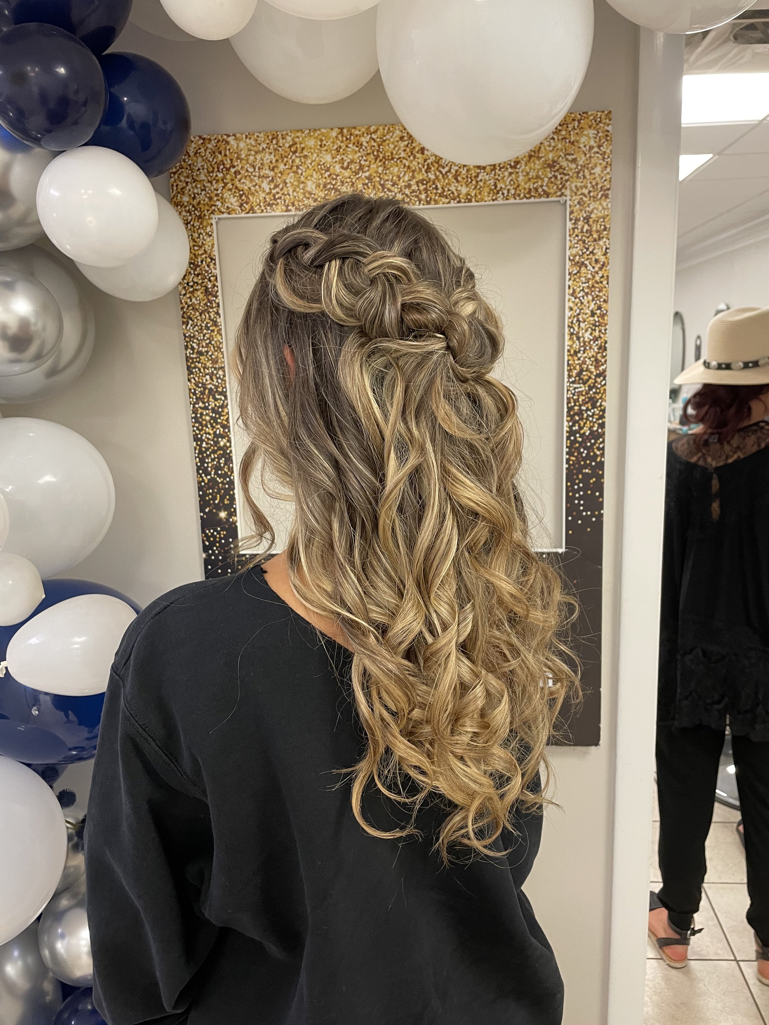 A woman with long, curly hair styled in an intricate braid, standing in front of a mirror with a gold and black glittery border, surrounded by white, navy blue, and silver balloons. In the background, a person is wearing a white hat and black dress.