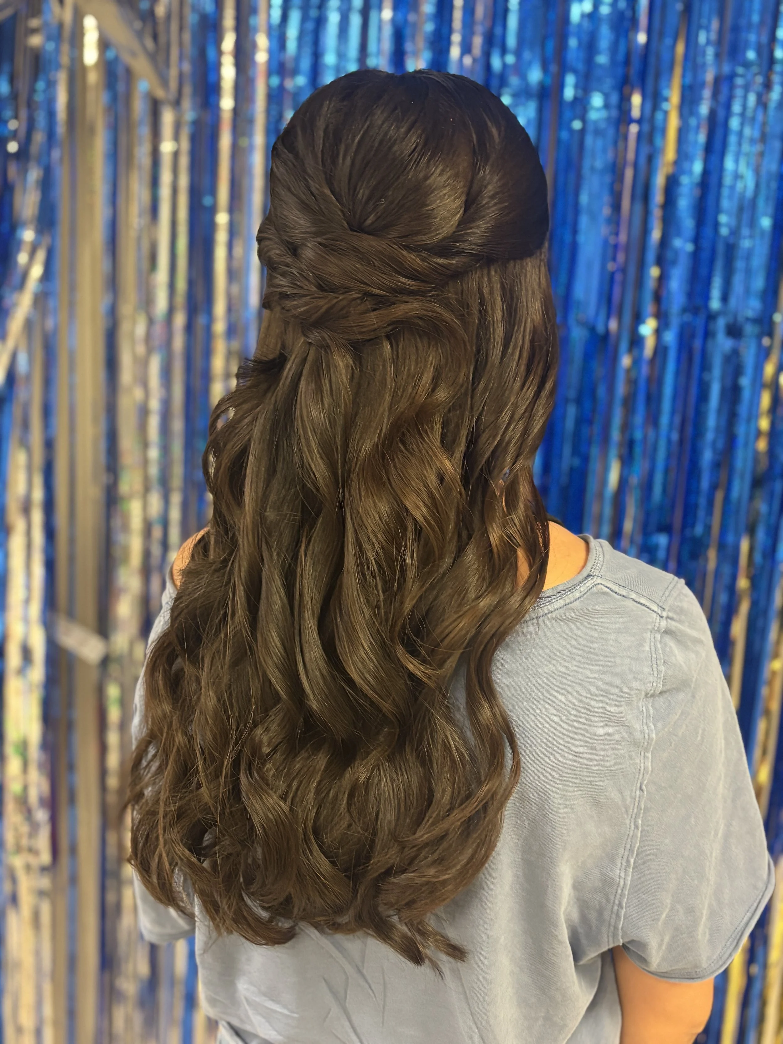 Back of a woman with long, wavy, brown hair styled in a loose braid, wearing a gray T-shirt, standing in front of a background of shiny blue metallic curtains.