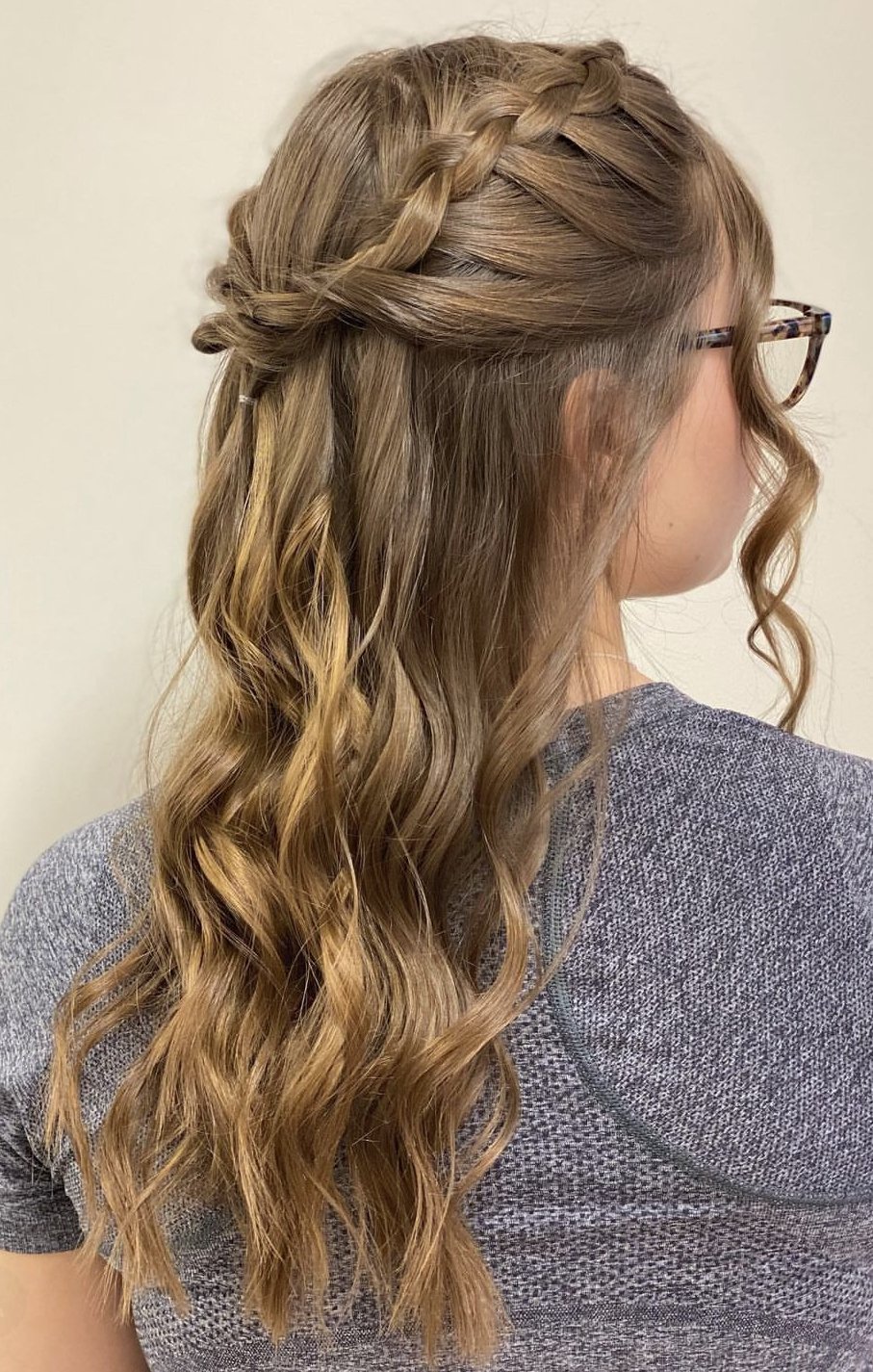 Side view of a woman with long, wavy, blonde hair styled in a loose braid crown, wearing glasses and a gray textured shirt.