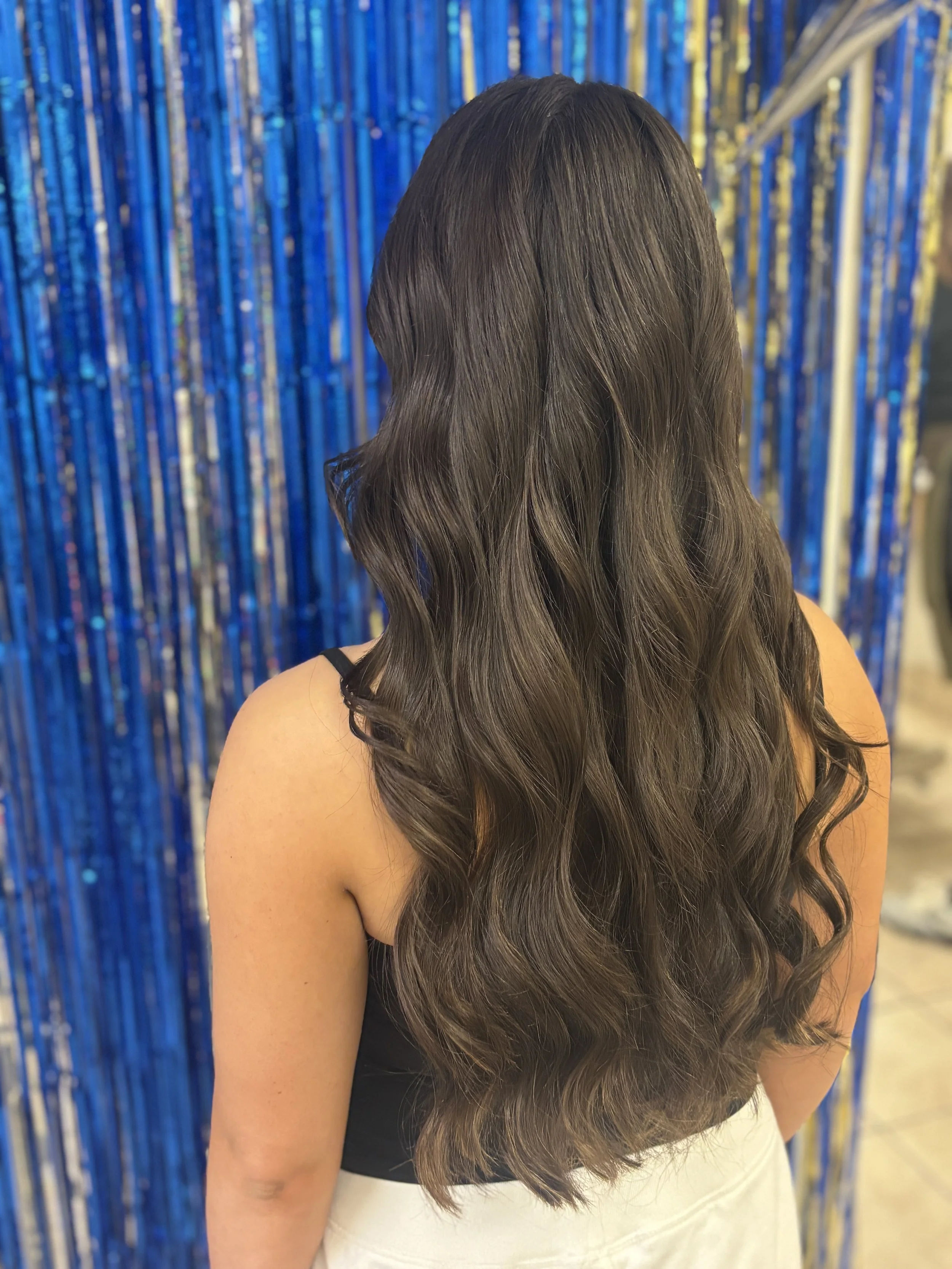 Back view of a woman with long, wavy brown hair standing in front of a blue metallic curtain.