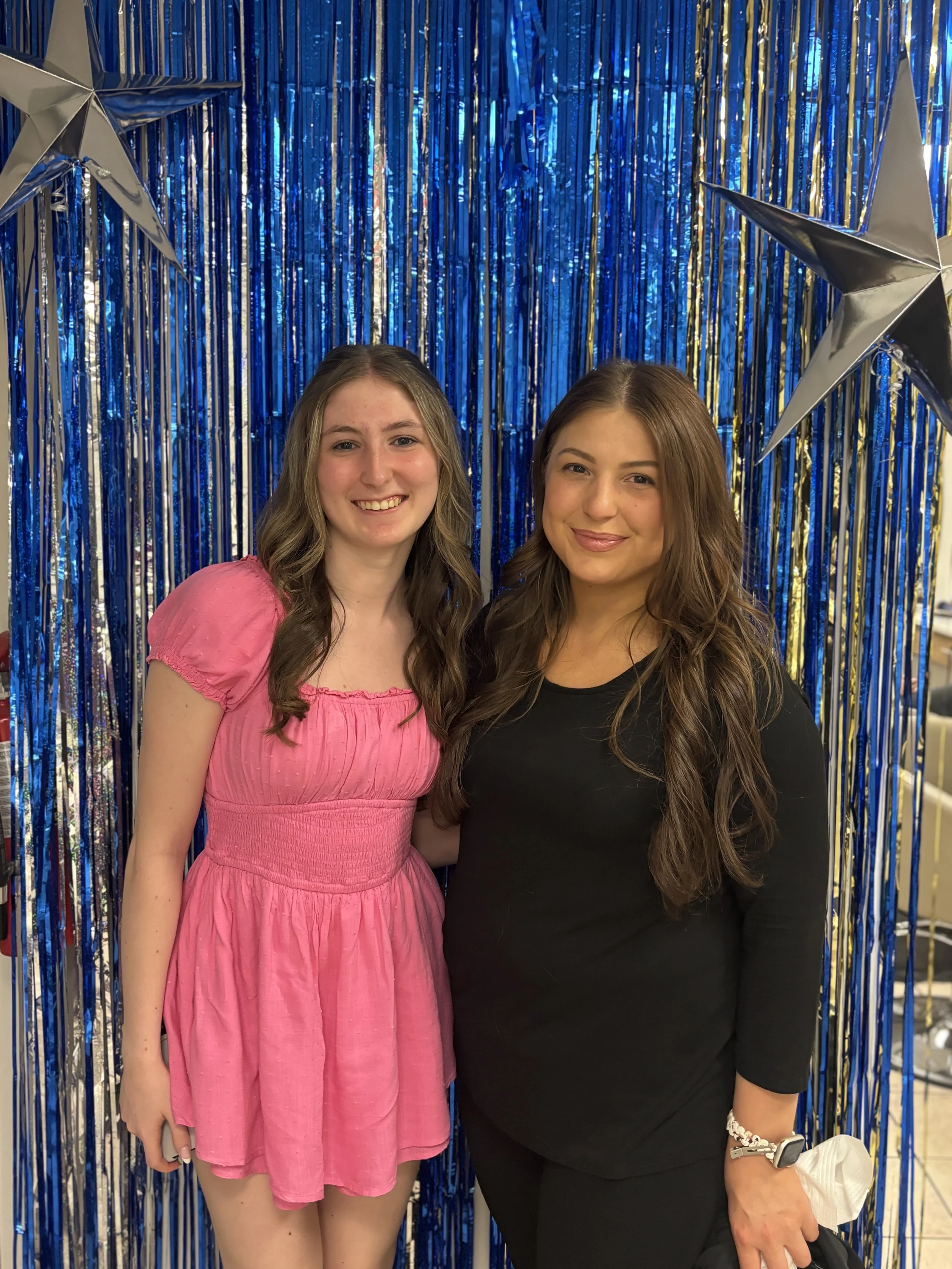 Two women standing in front of a blue metallic curtain with large silver star decorations, smiling at the camera.