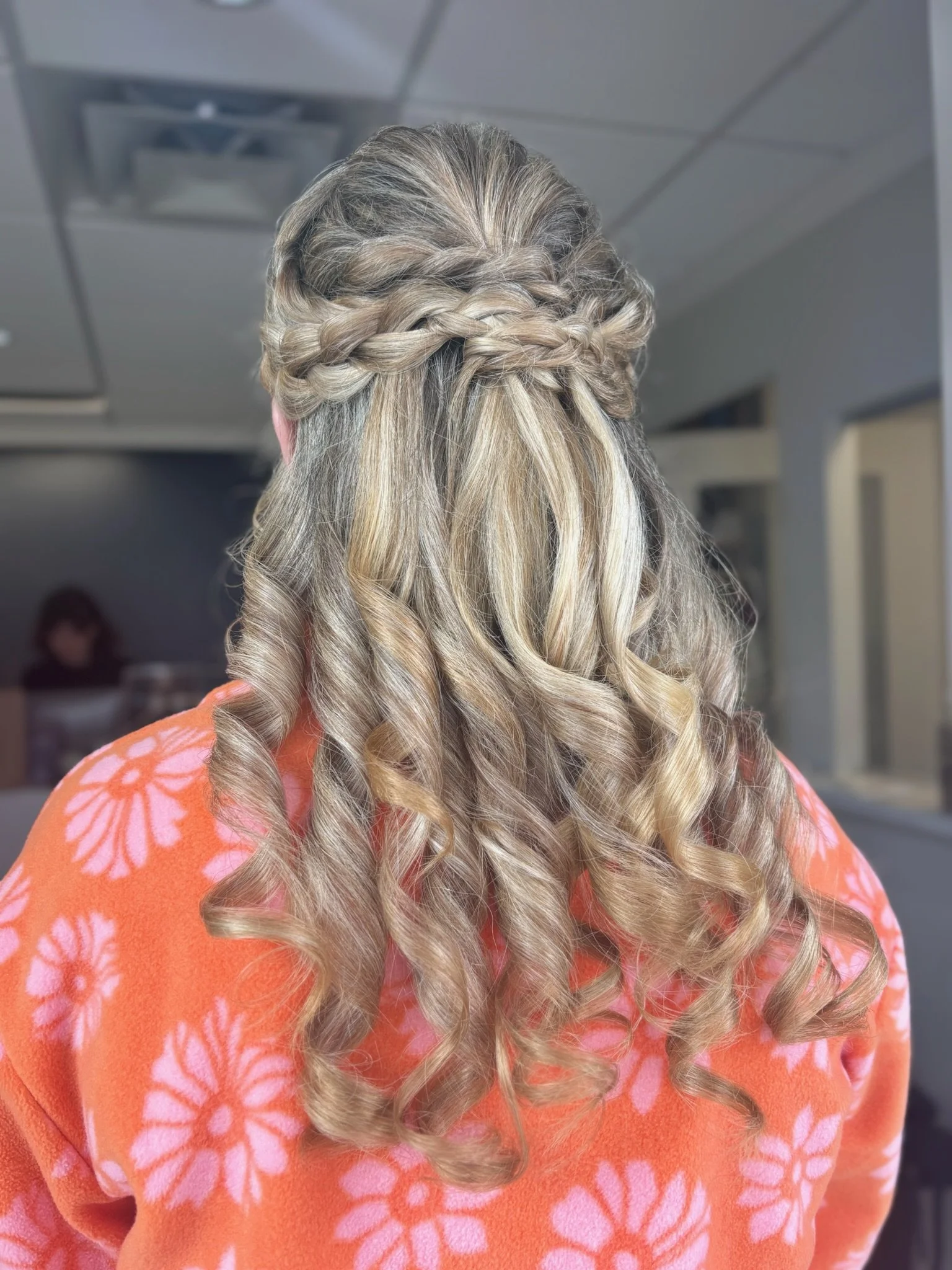 Back view of a woman with long, curly blonde hair styled in a braided half-updo, wearing an orange garment with pink floral patterns.