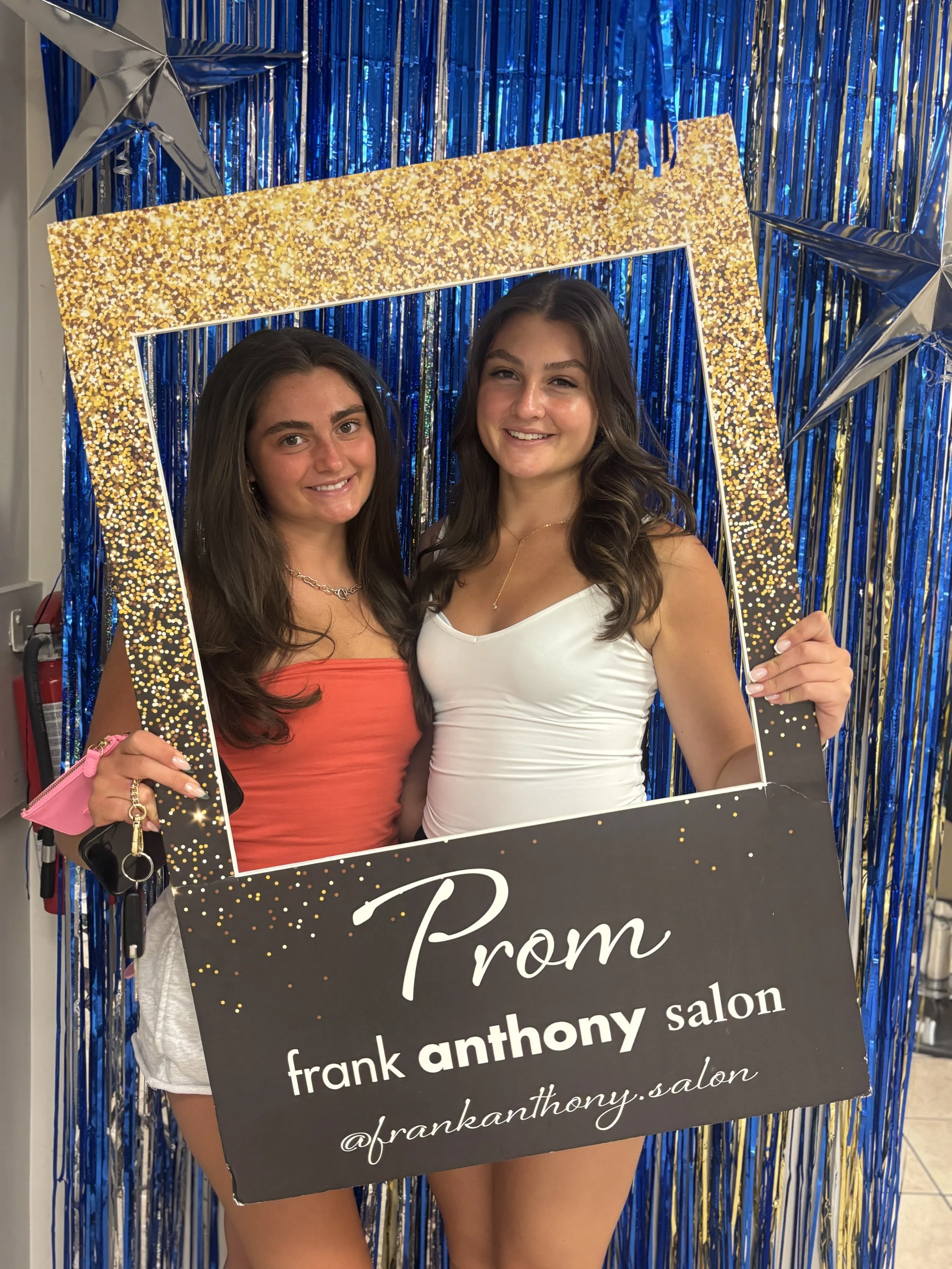 Two young women smiling and holding a sign that reads 'Prom' at a celebration event with a blue and gold tinsel backdrop.