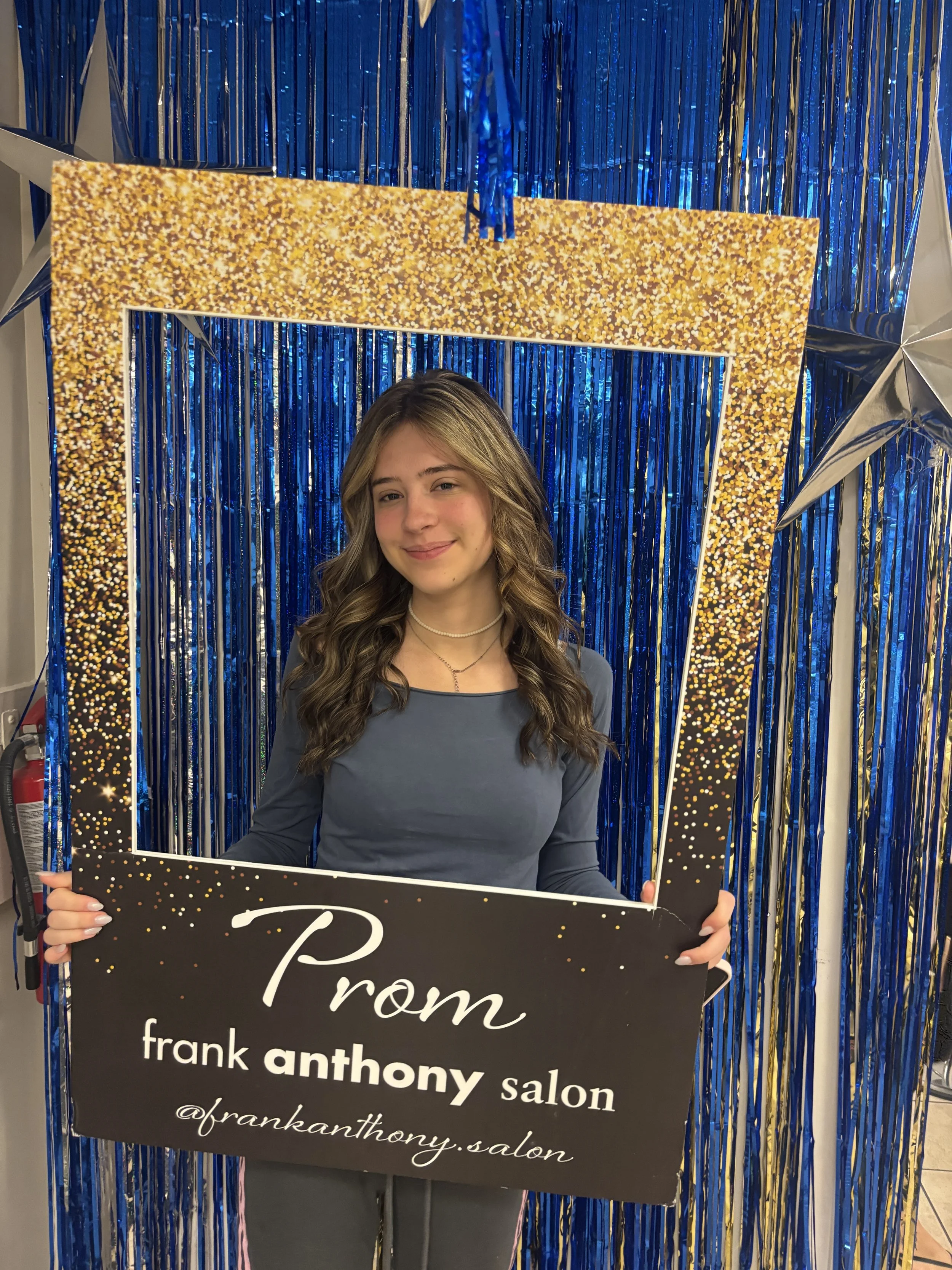 A young woman with long, wavy hair and a gray top holding a black sign with 'Prom' written on it, standing in front of blue metallic streamers and silver star decorations at a prom event.