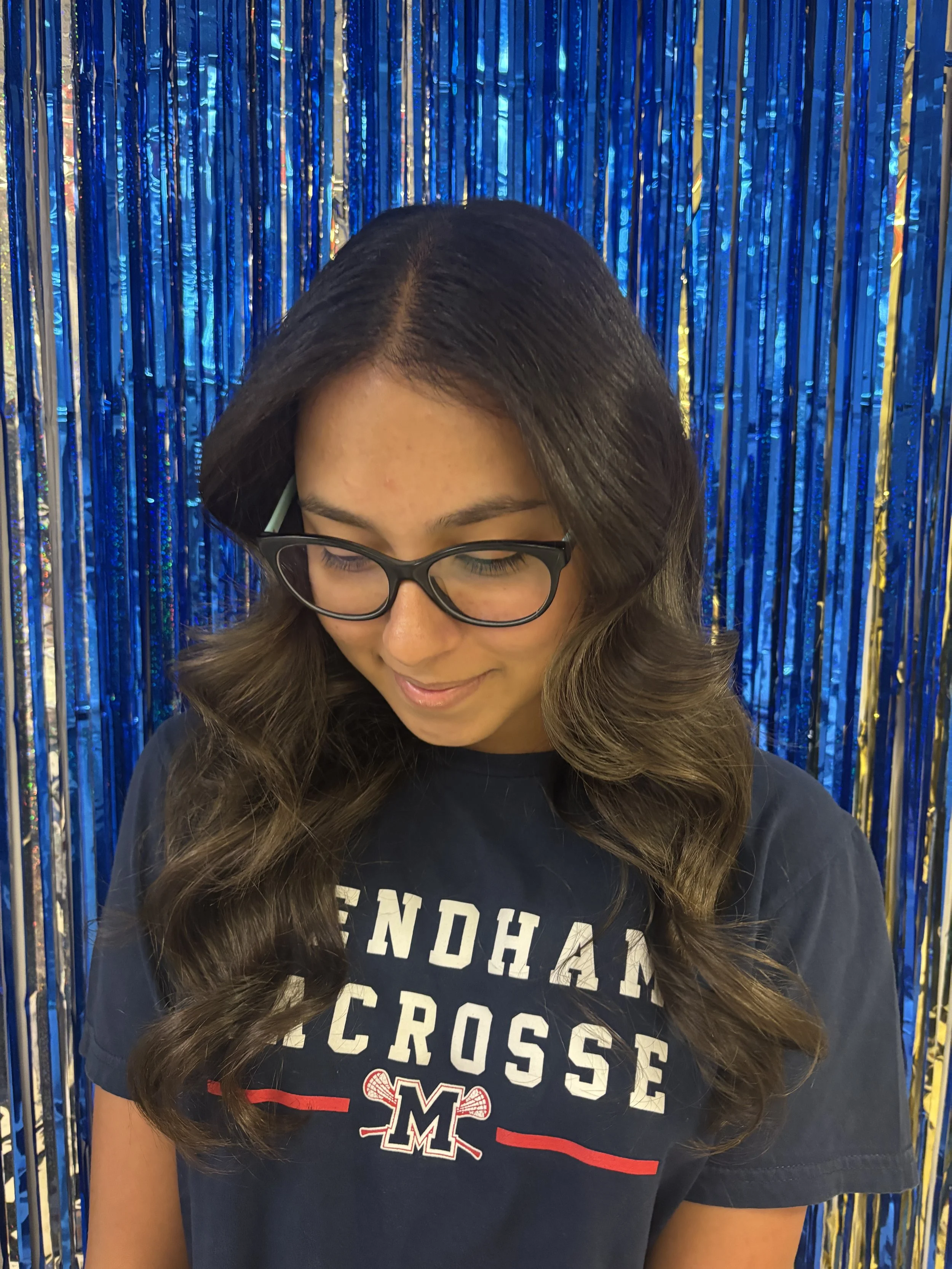 A young woman with long, wavy brunette hair, wearing glasses and a navy blue sports shirt with 'CROSSFIT' written on it, standing in front of a shiny blue and gold metallic curtain.