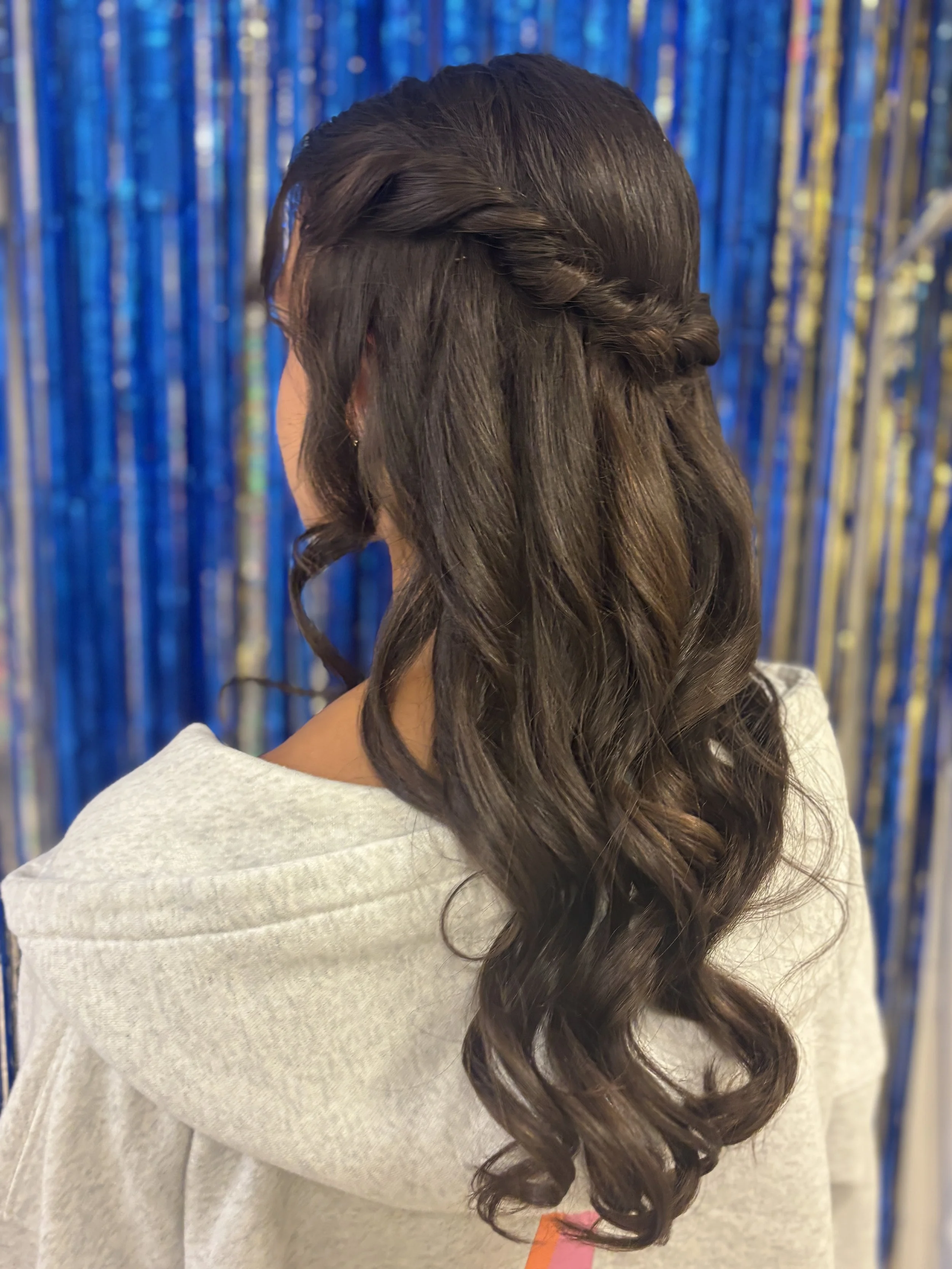 Back view of a woman with long, wavy brown hair styled in a twisted half-updo, standing in front of a blue shimmering curtain.
