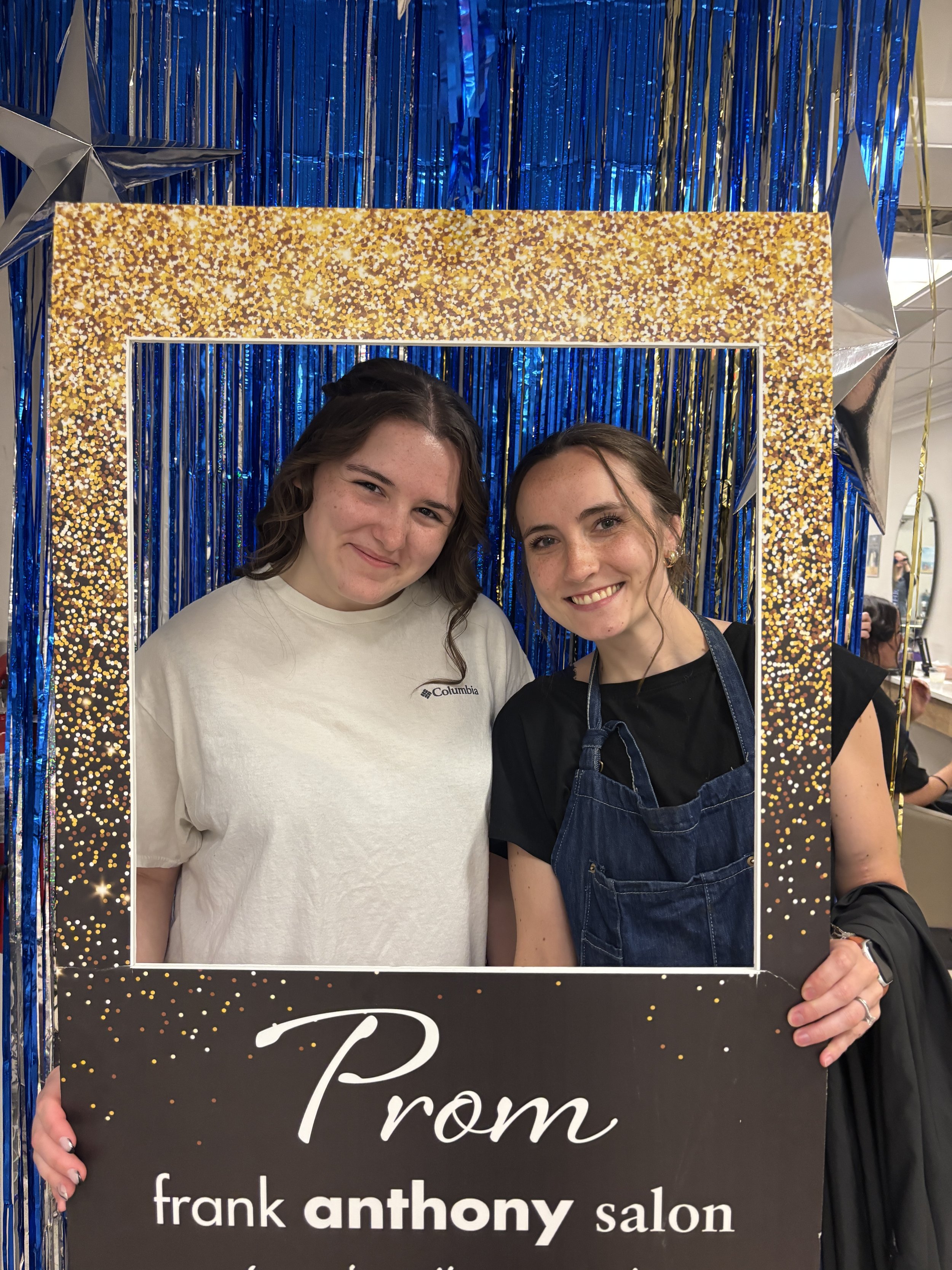 Two young women standing behind a large black and gold sign with the words 'From frank anthony salon'. They are smiling and partially behind a decorative frame, with a blue and gold festive backdrop behind them.