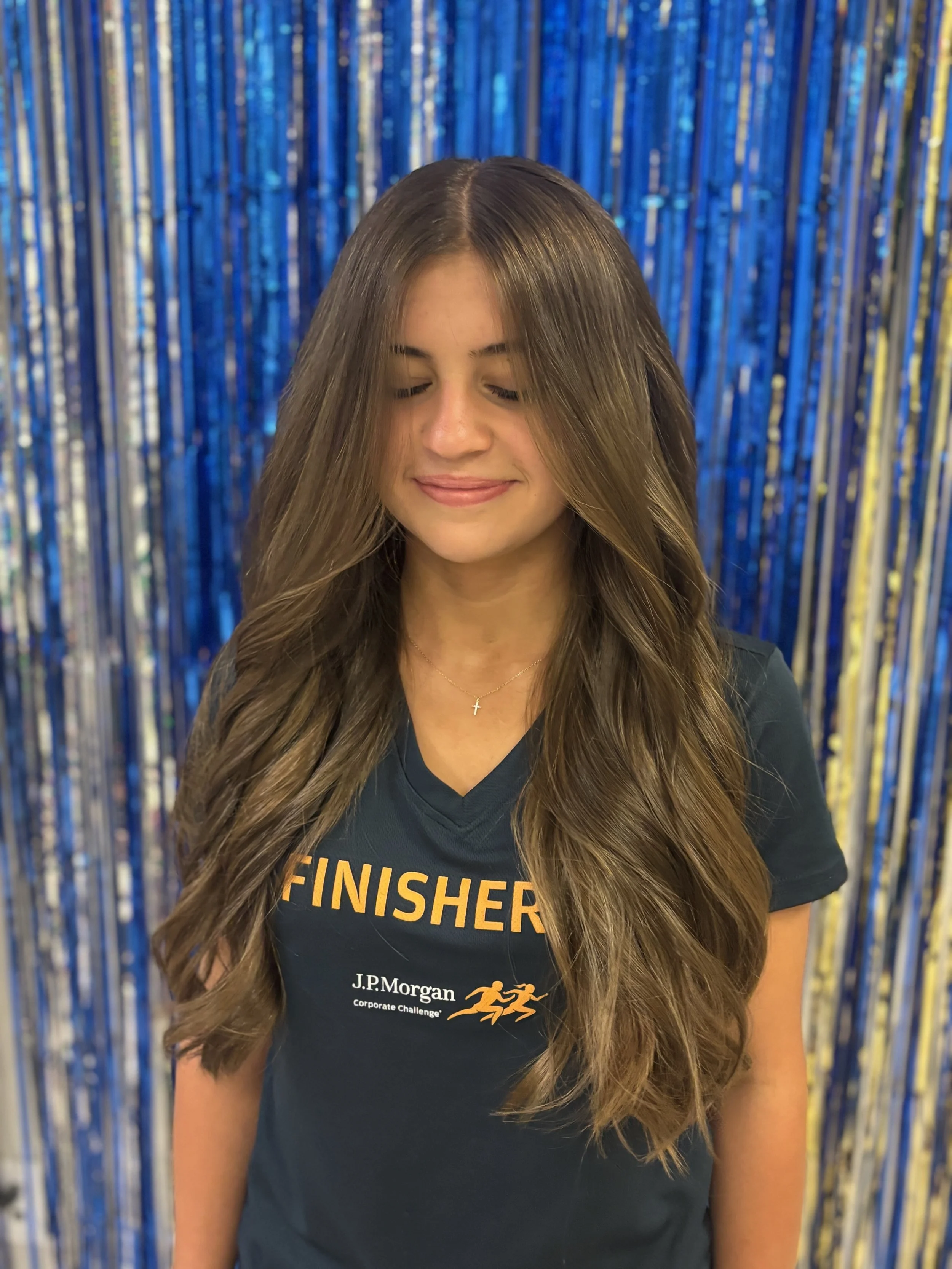 A young woman with long, wavy brown hair and closed eyes, smiling softly, wearing a dark blue shirt with orange text that says 'FINISHED' and additional logo and text below, standing in front of a blue and gold metallic streamer backdrop.