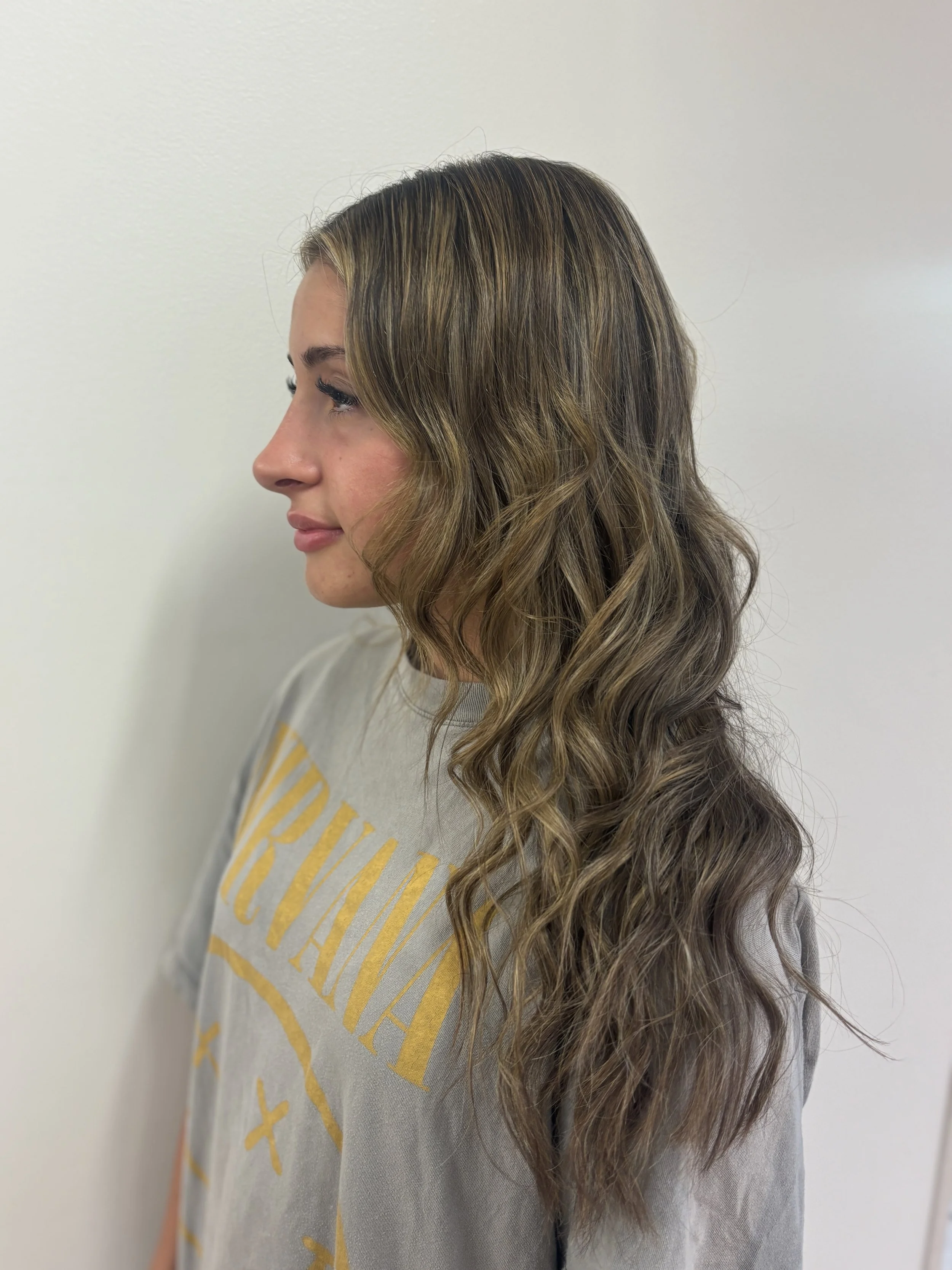 Profile of a woman with long, wavy hair styled in loose curls, wearing a gray shirt with yellow text, standing against a plain white wall.