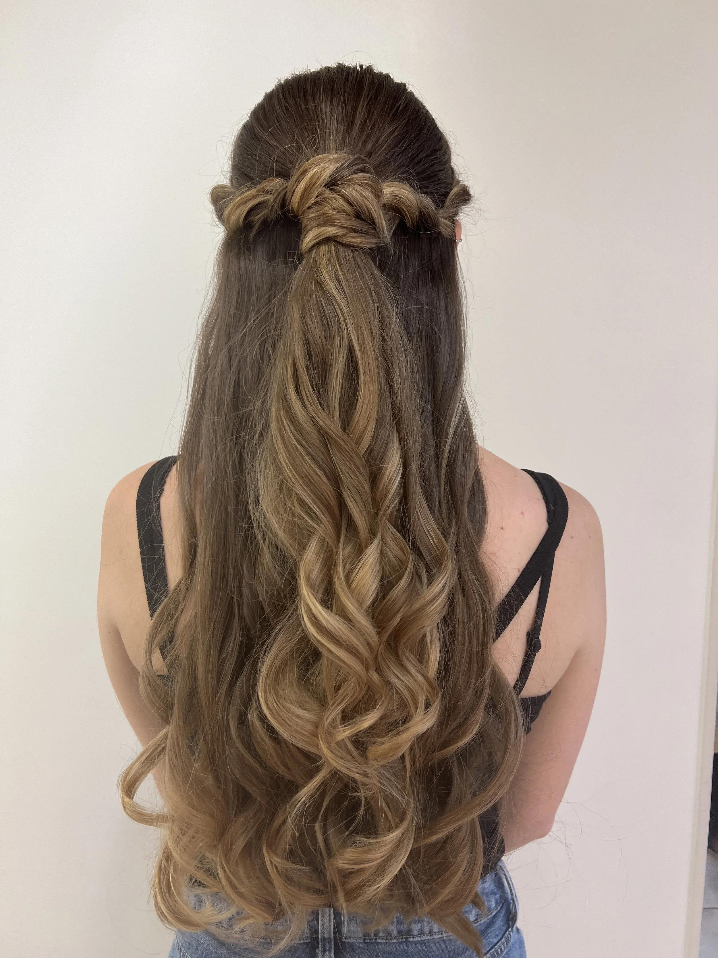 A person with long, wavy, ombre hair styled in a half-up, twisted braid, wearing a black tank top and blue jeans, standing against a plain white background.