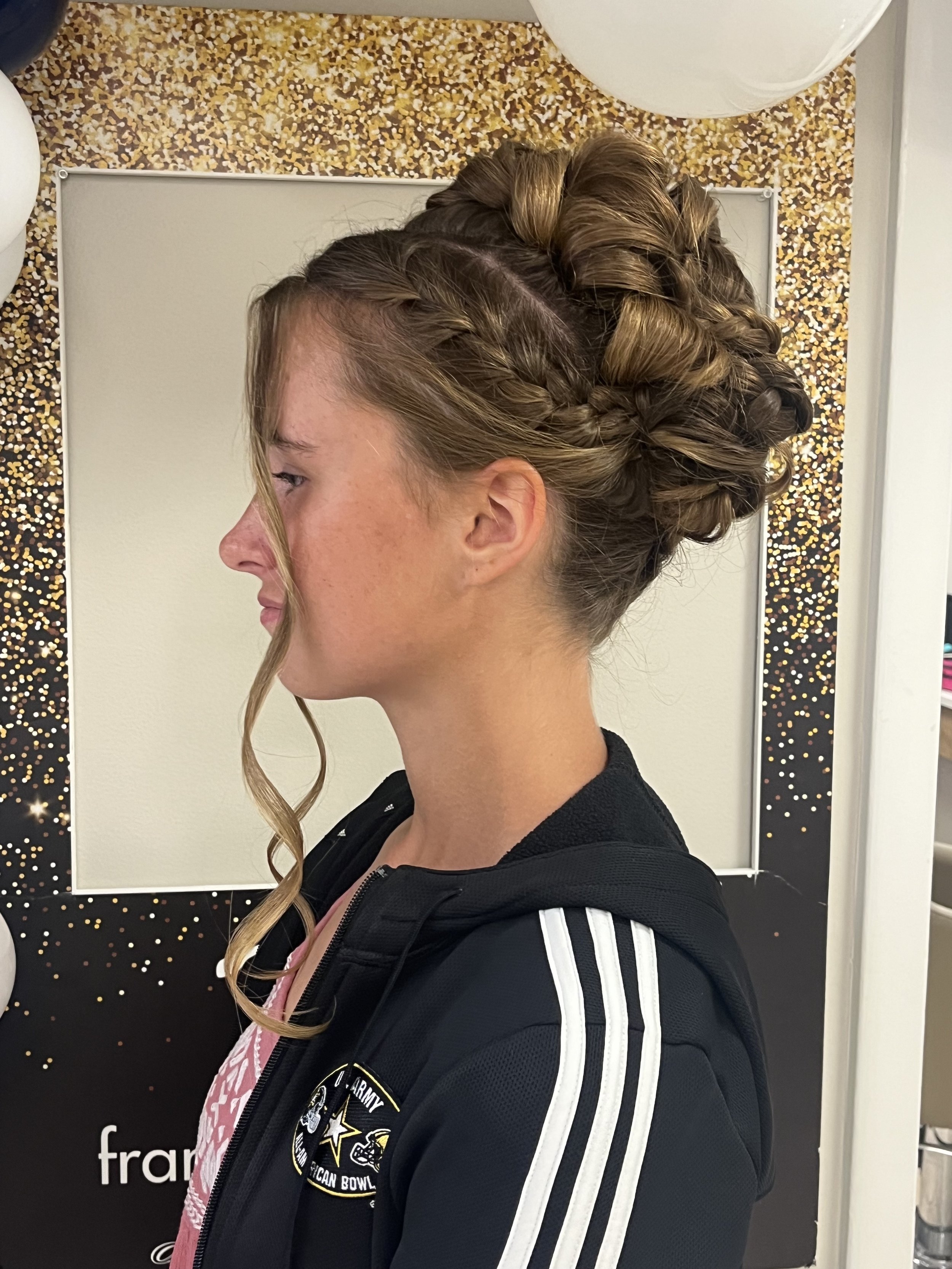 Side profile of a woman with her hair styled in an elegant updo with braided and rolled sections, standing in front of a mirror.