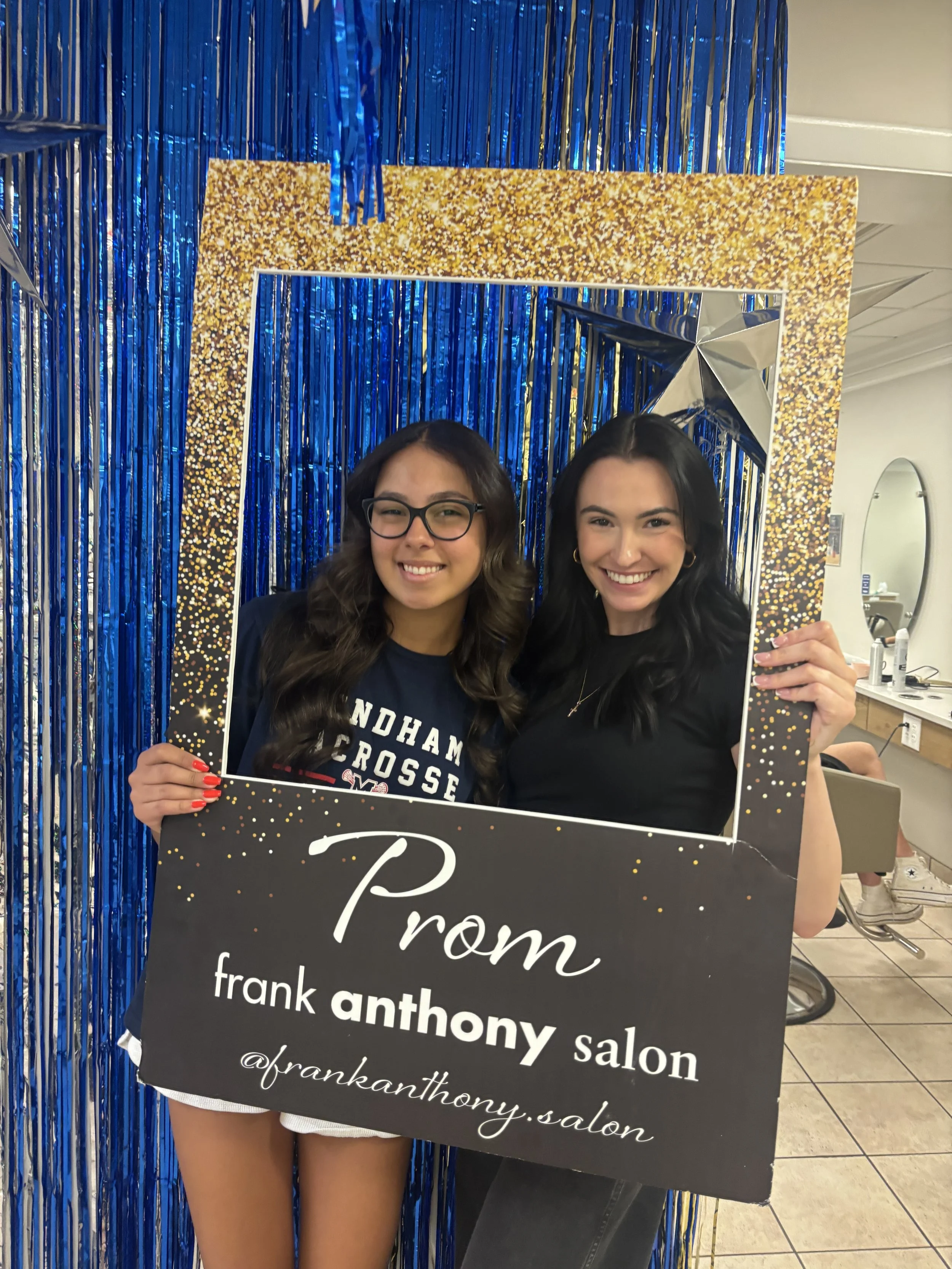 Two young women holding a black and gold sign that reads 'Prom from frank antony salon' inside a room decorated with blue tinsel and gold confetti accents, taking a selfie through a photo frame