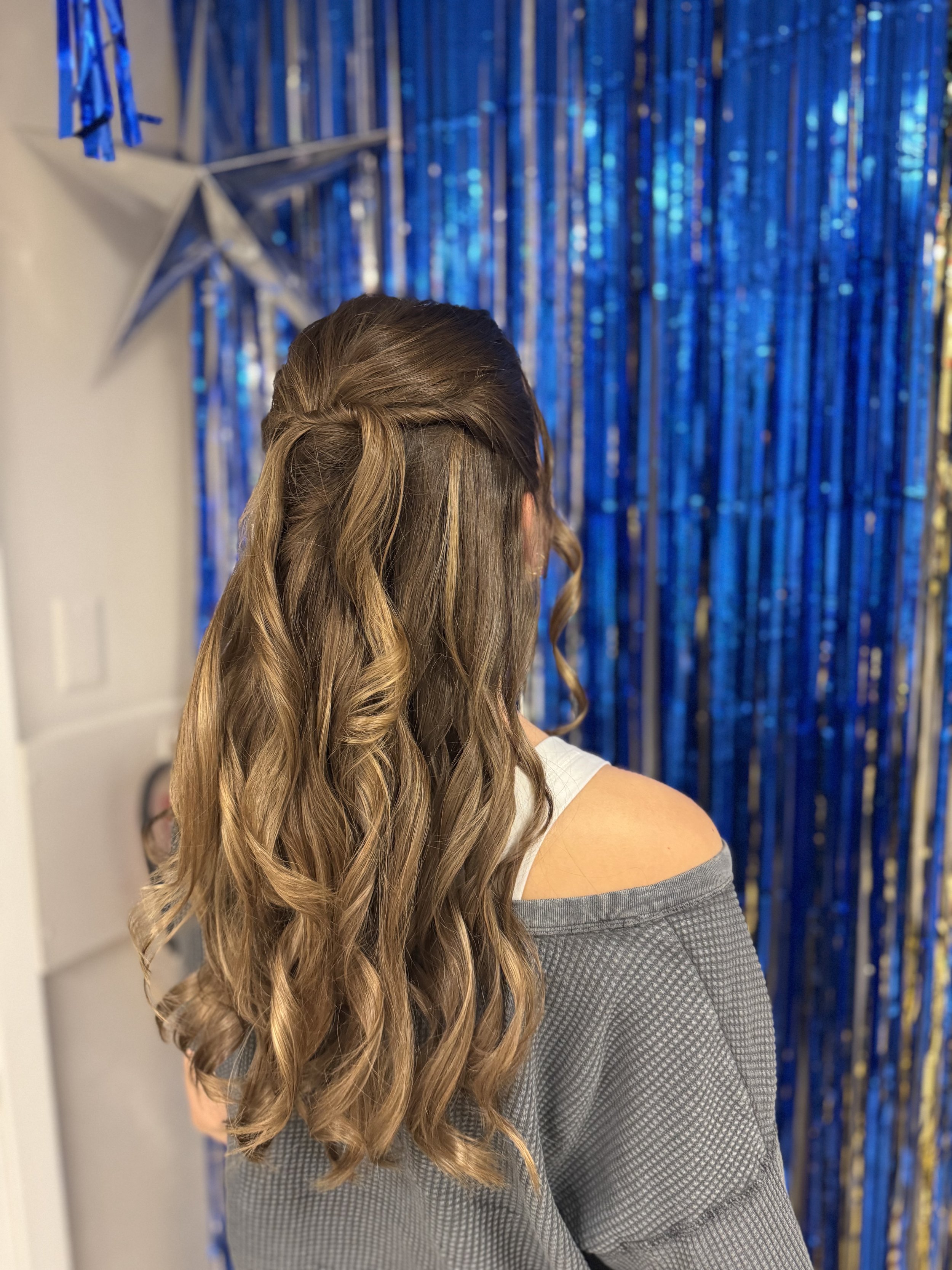 Back view of woman with long, curled, light brown hair wearing a gray cold shoulder top, in front of a shiny blue metallic curtain, some star-shaped decorations are on the left side.