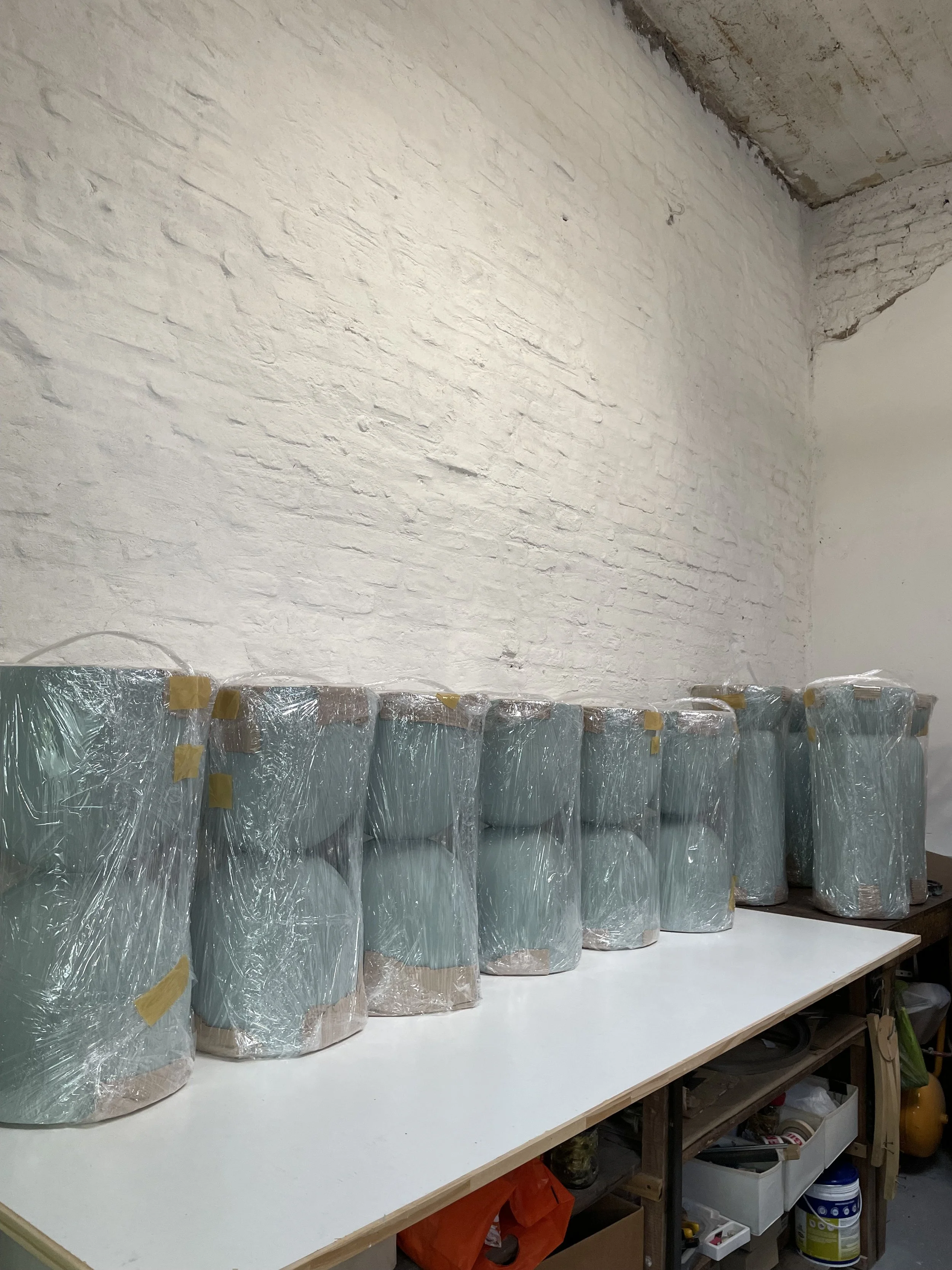 Eight wrapped ceramic vases or pots are lined up on a white table against a white brick wall in a workshop or storage room.