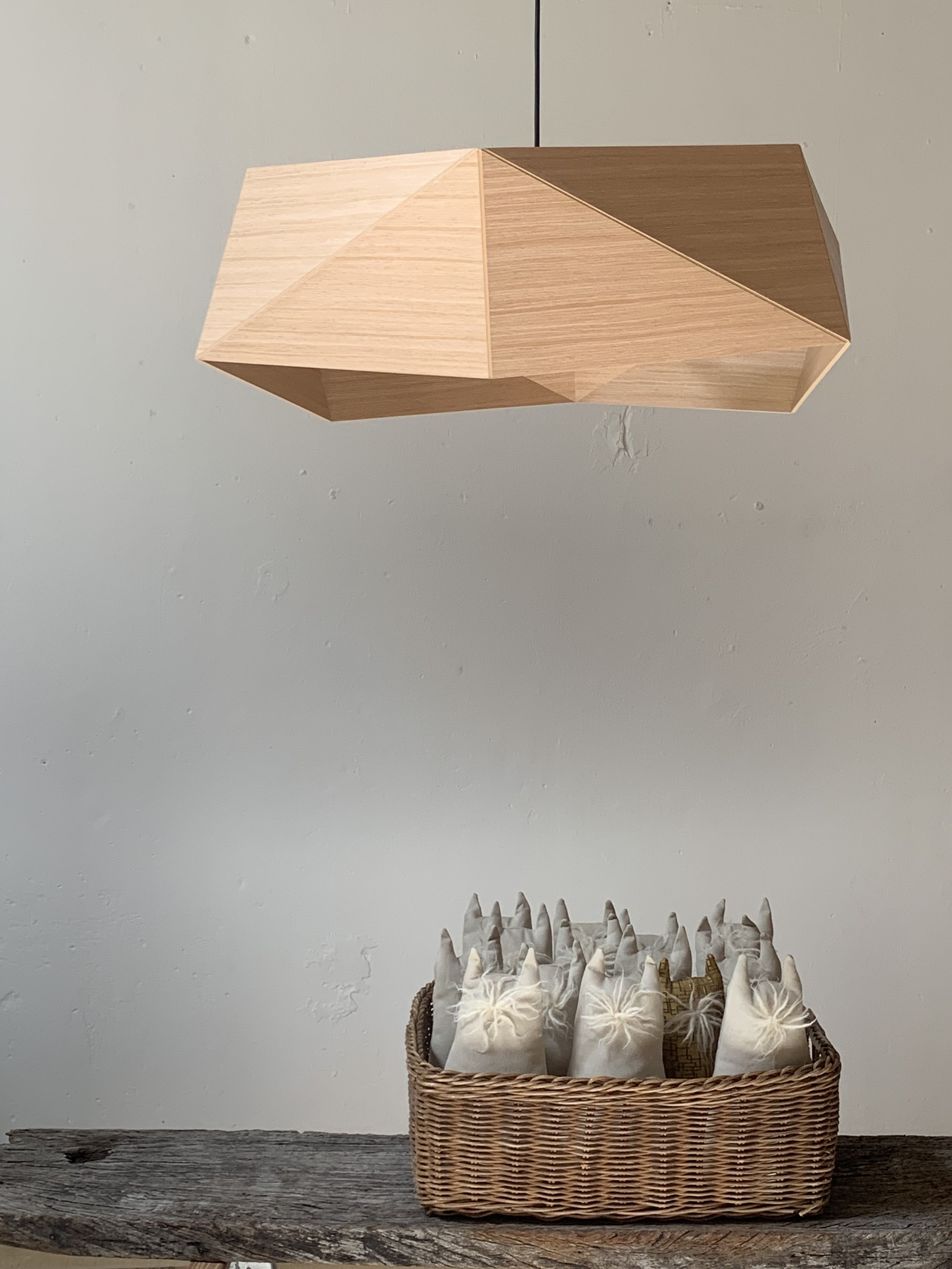 Wooden geometric pendant lamp hanging above a basket filled with fabric-wrapped items on a wooden surface.
