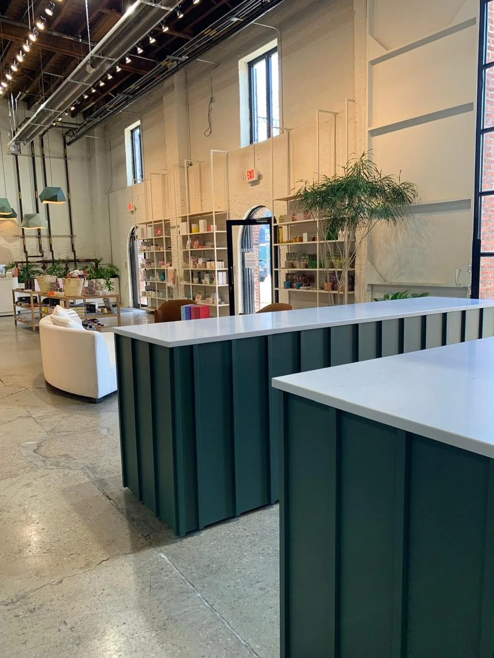 Modern interior of a bookstore or library with high ceilings, white walls, large windows, bookshelves, green and white tables, seating area with a white couch, and plants.