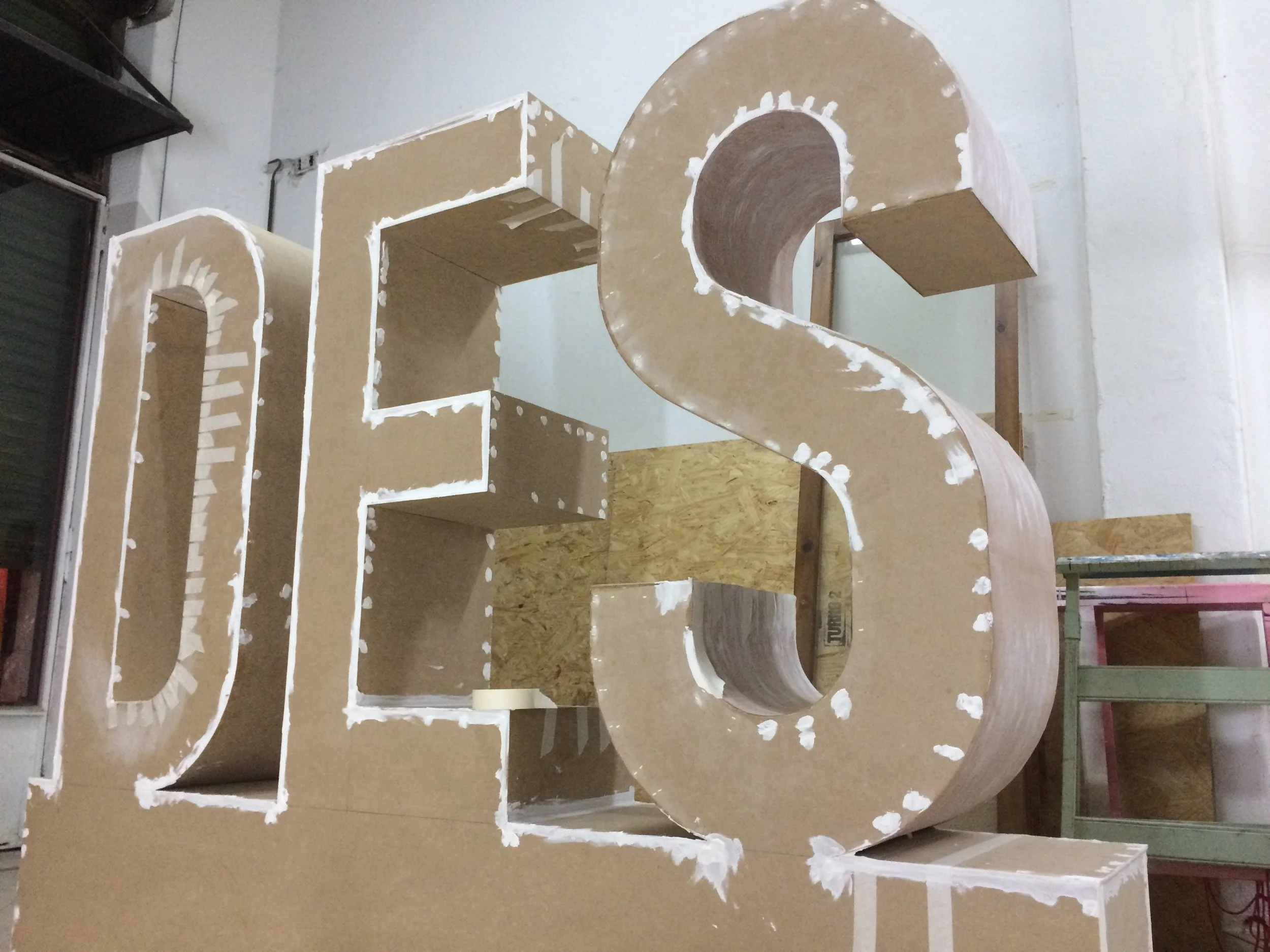 A large, three-dimensional letter 'S' made of wood and cardboard, with white paint or glue around the edges, positioned on a worktable in a workshop.