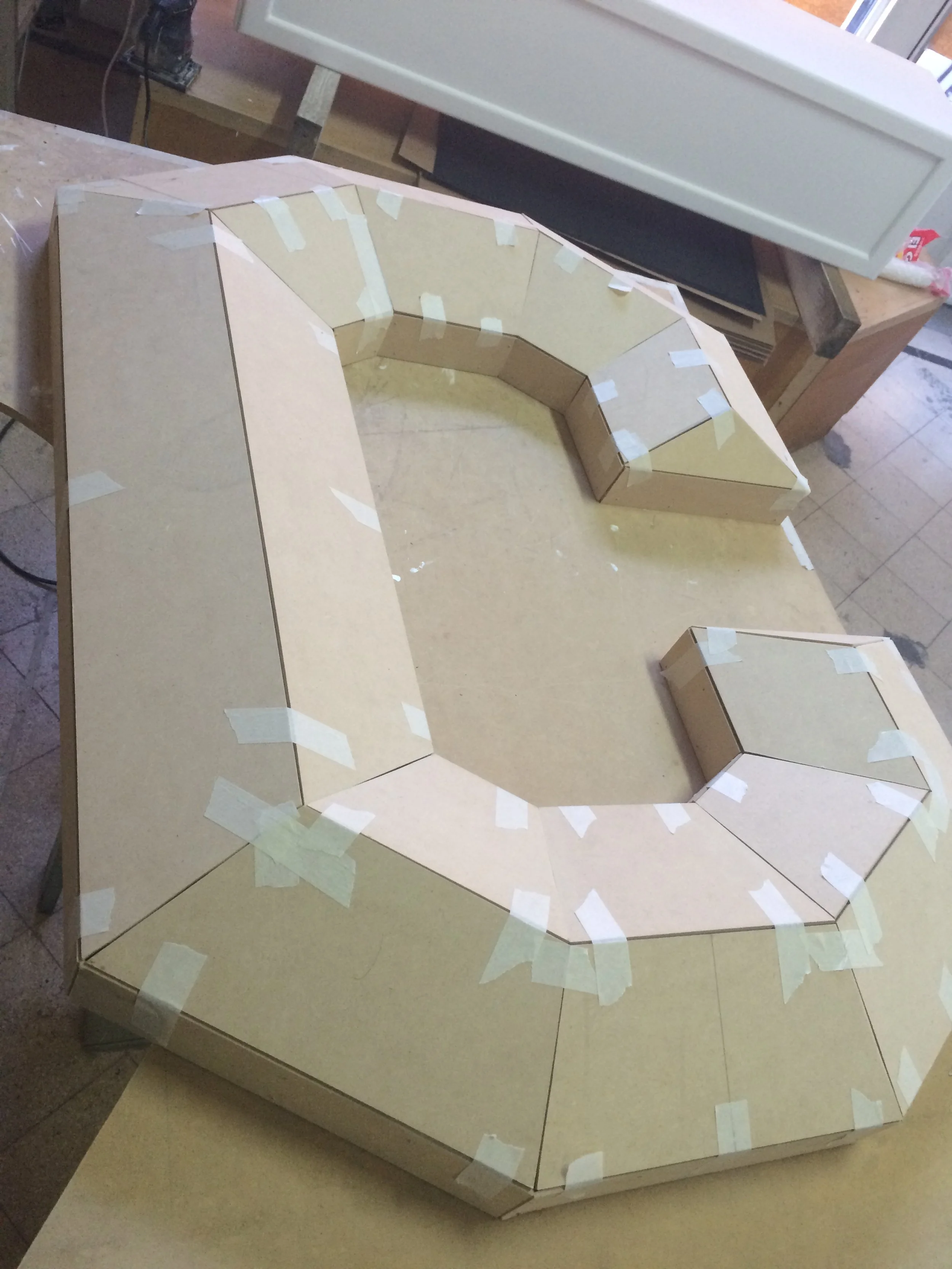 Unfinished cardboard model of a circular structure with multiple angled panels, taped together, on a wooden work surface in a workshop.