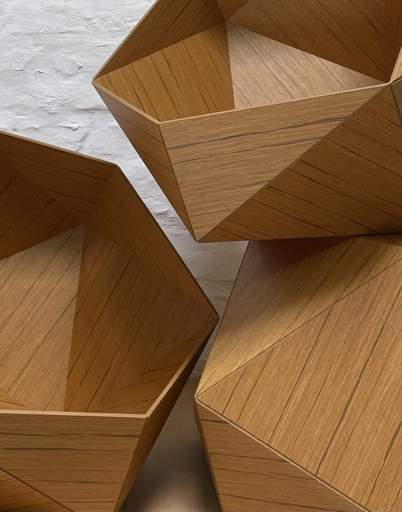 Three wooden geometric open-top containers with angled sides and visible wood grain, placed against a white textured wall.