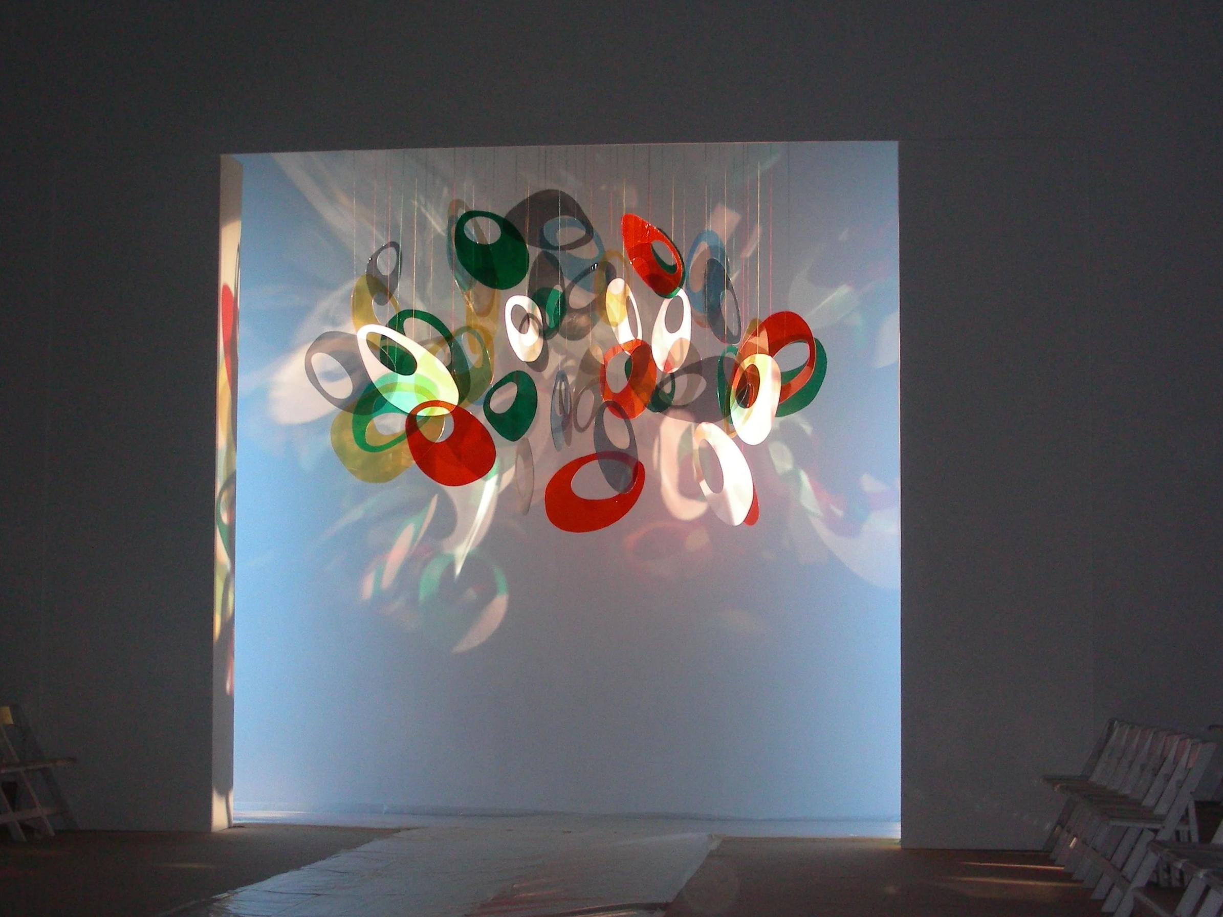 An art installation of colorful oval-shaped discs suspended from the ceiling in an indoor space with chairs along the right wall.
