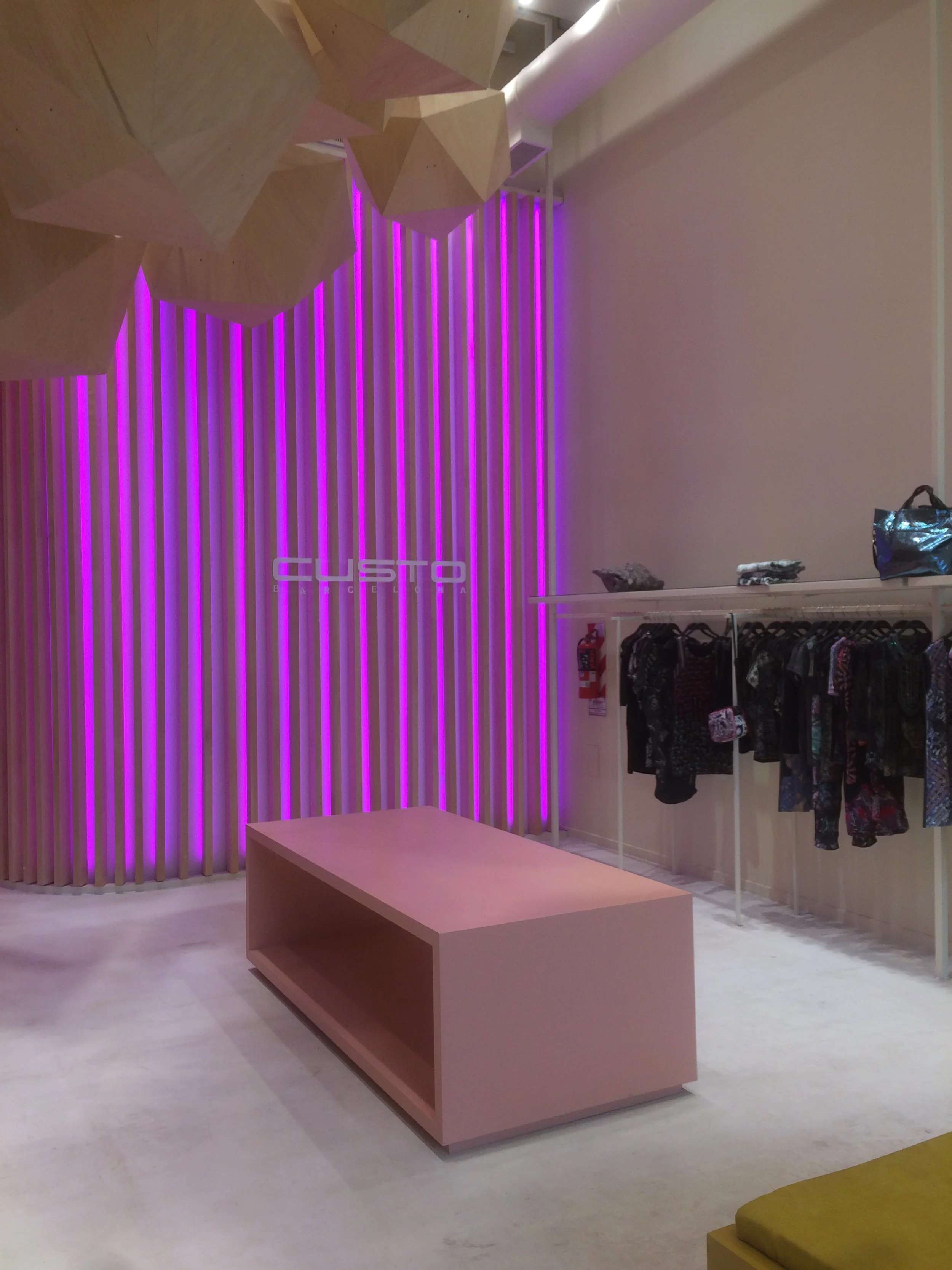 Interior view of a boutique store with pink for display table, purple neon lights, clothing on racks, and geometric wooden lighting fixtures on the ceiling.