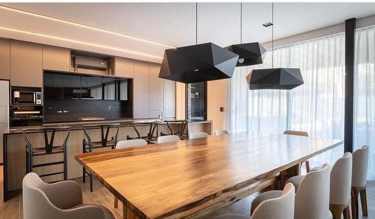Modern dining room with a large wooden table, white upholstered chairs, a kitchen counter with barstools, and geometric black pendant lights. Large windows with sheer curtains let in natural light.