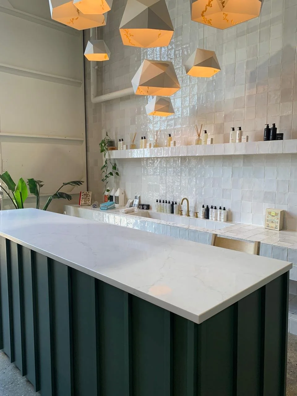 Modern interior space with a white marble countertop island, beige tiled backsplash, decorative geometric pendant lights, and shelves with skincare or cosmetic bottles and containers, accented with green plants.