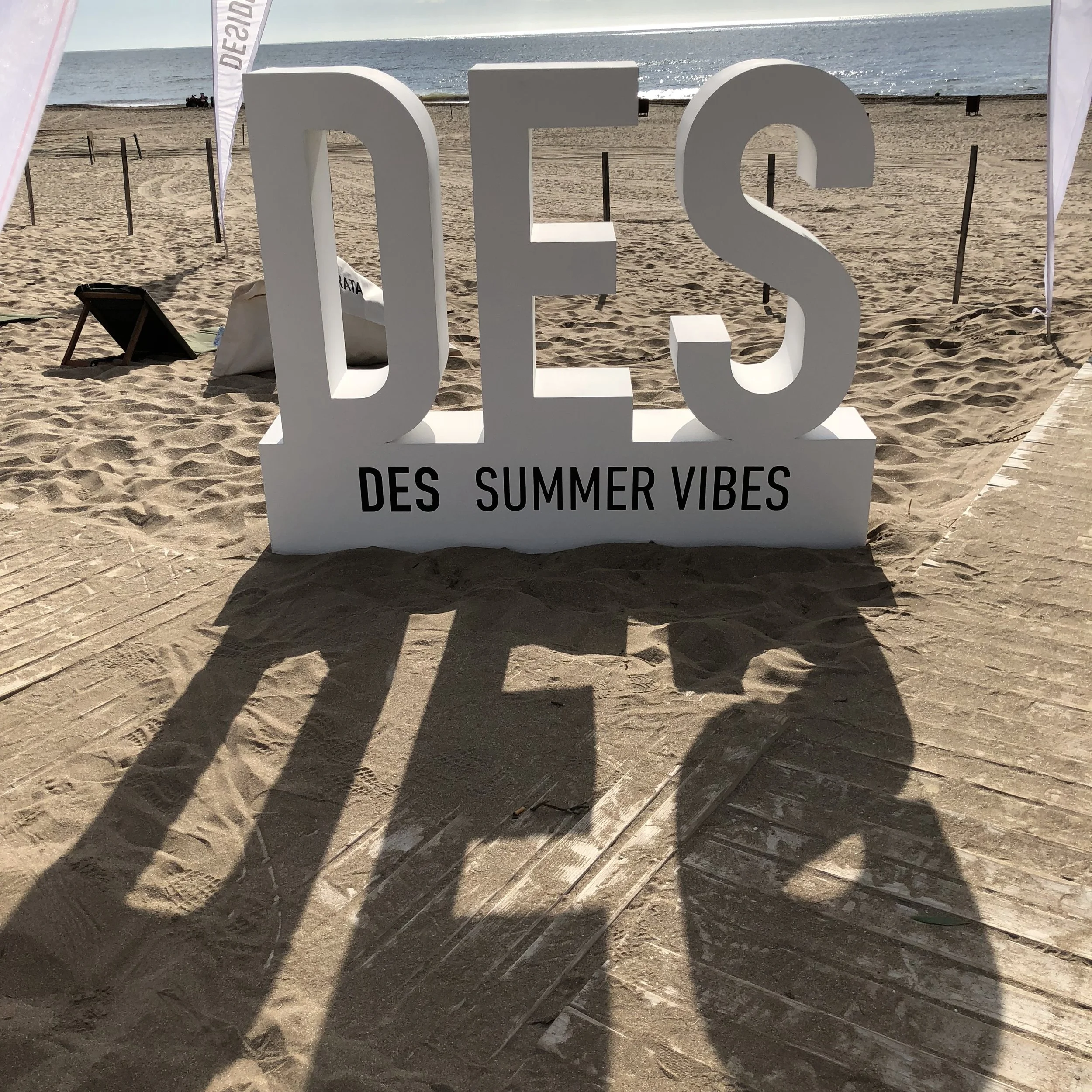 Large white sign on the sandy beach spelling out 'DES' with shadows cast on the sand, and smaller text reading 'DES Summer Vibes' below.