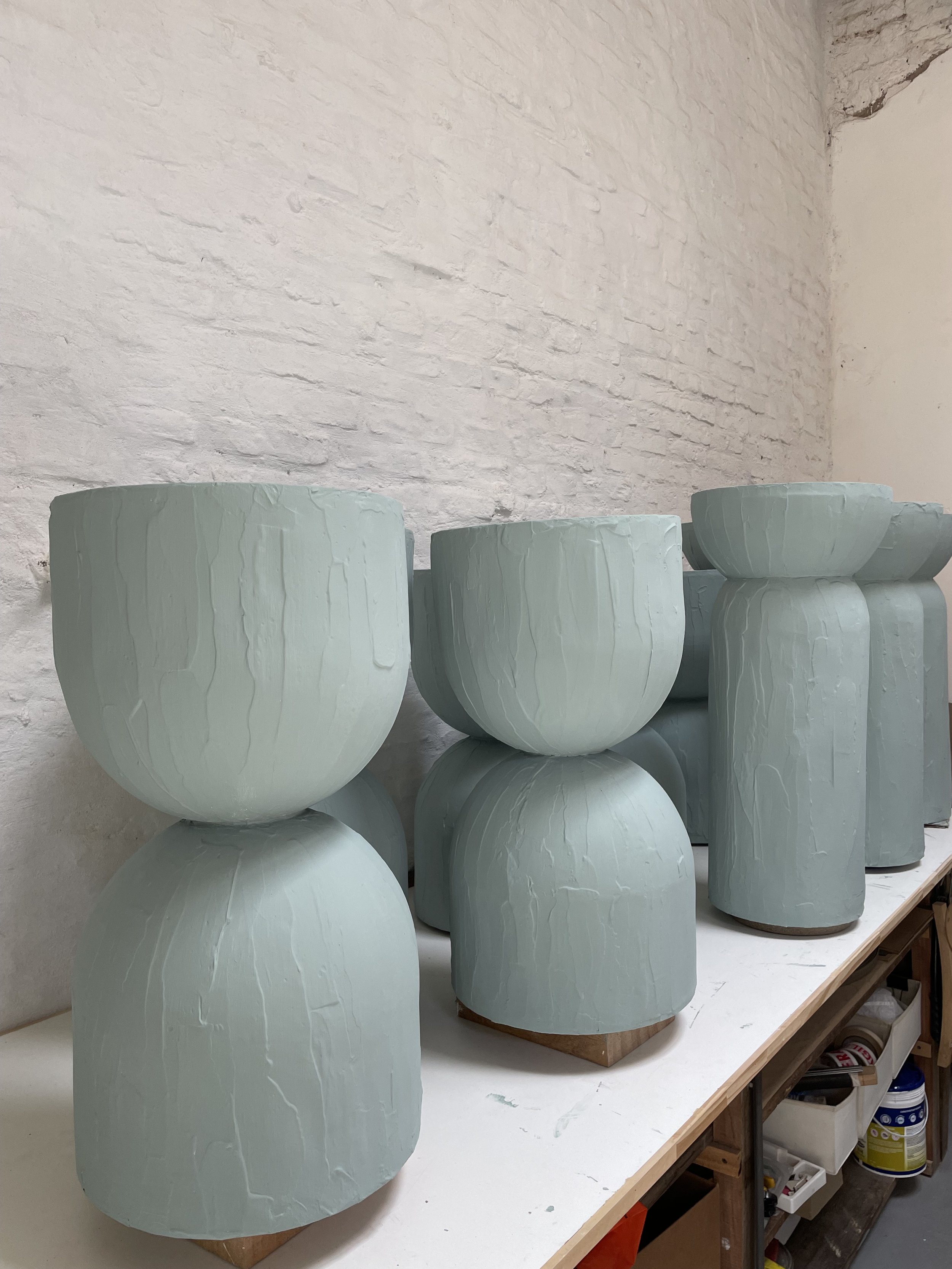 Several large, light green ceramic vases with textured surfaces, placed on a workbench against a white brick wall.