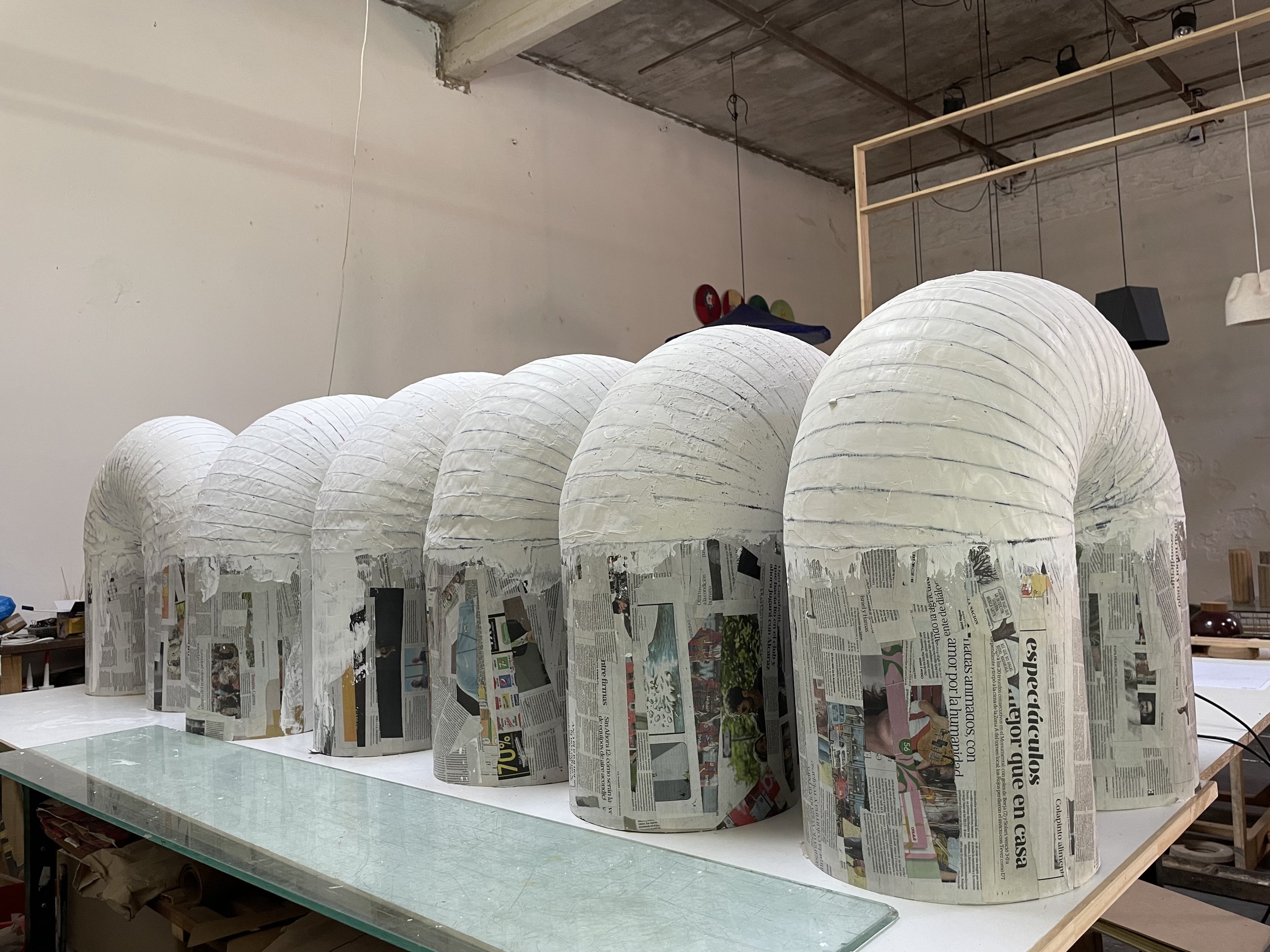 Several large, white, domed paper lanterns covered in newspaper are lined up on a table in a workshop setting.