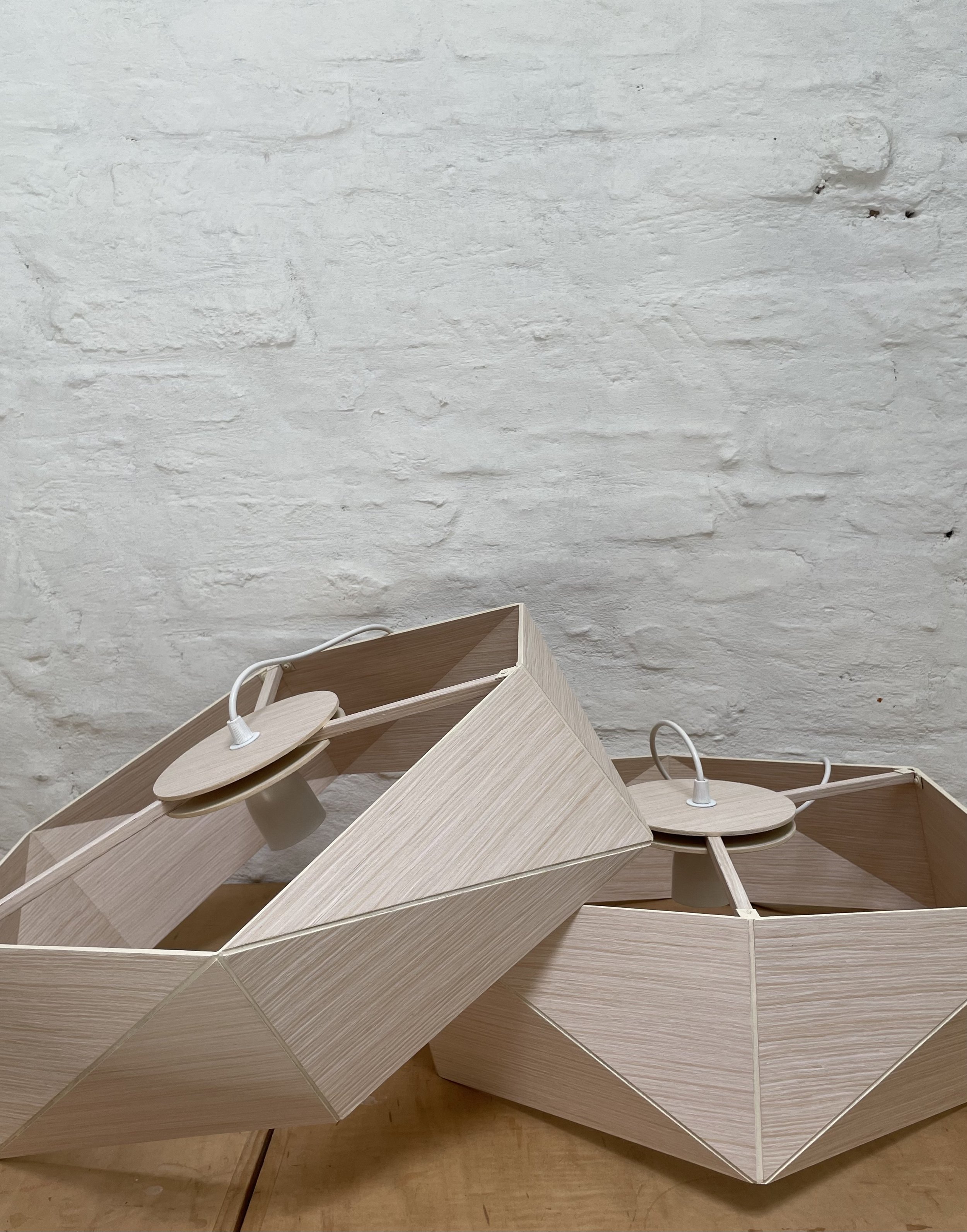 Two wooden geometric-shaped lamps with white electrical cords on a wooden surface against a white brick wall.